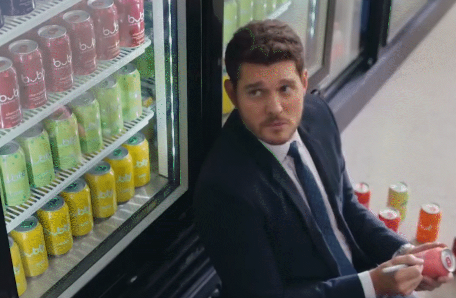 Man in a suit holding a beverage can beside a refrigerated display stocked with colorful soda cans