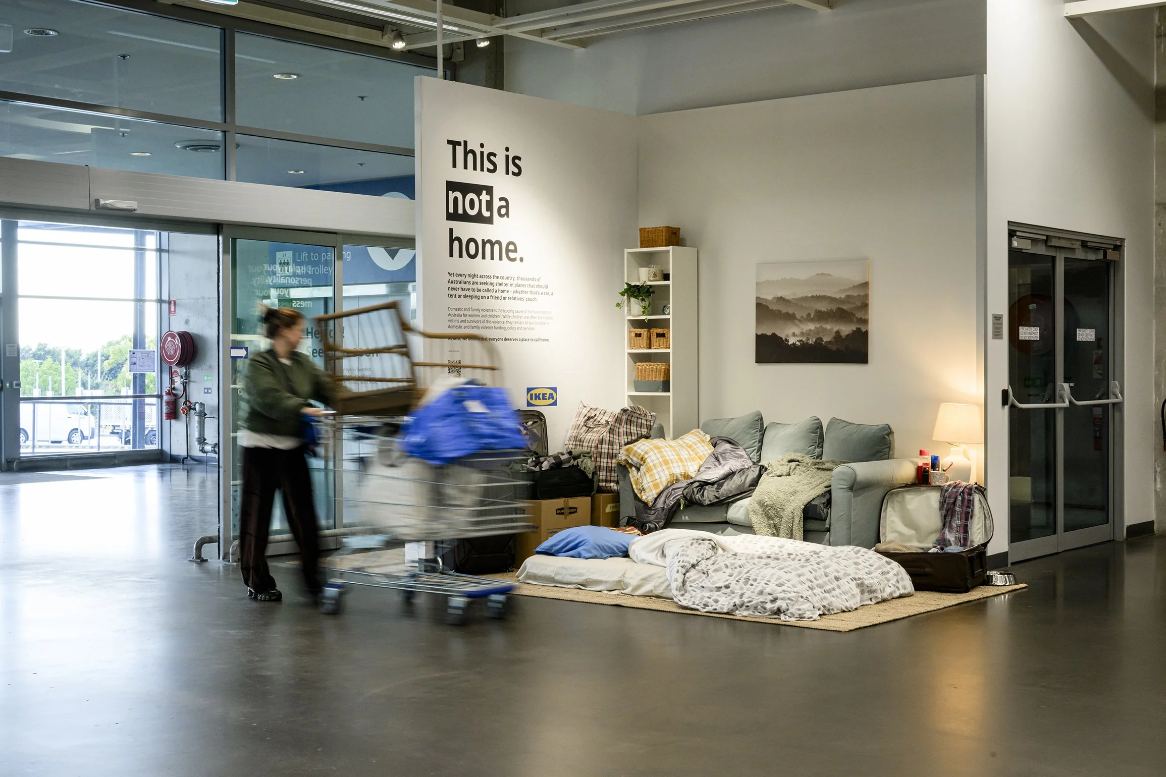 IKEA store display recreating a temporary home with a sofa, bedding, luggage, lamp, and advocacy sign reading “This is not a home.”