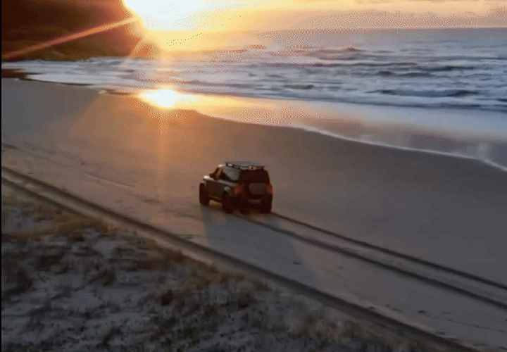 Small remote-controlled truck driving along a beach at sunset beside the ocean