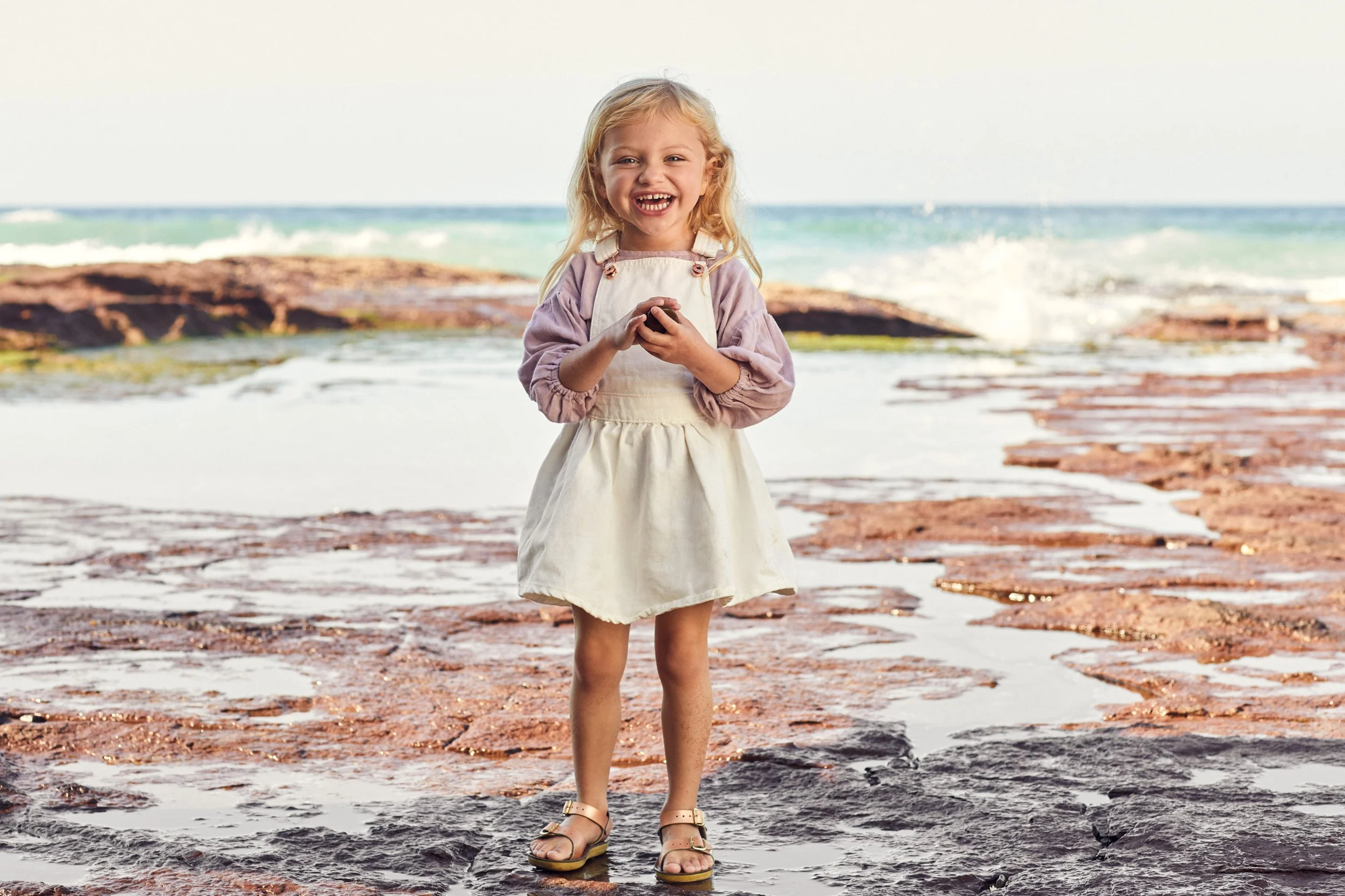 Smiling young girl in a white dress standing on rocky shore with ocean waves behind her