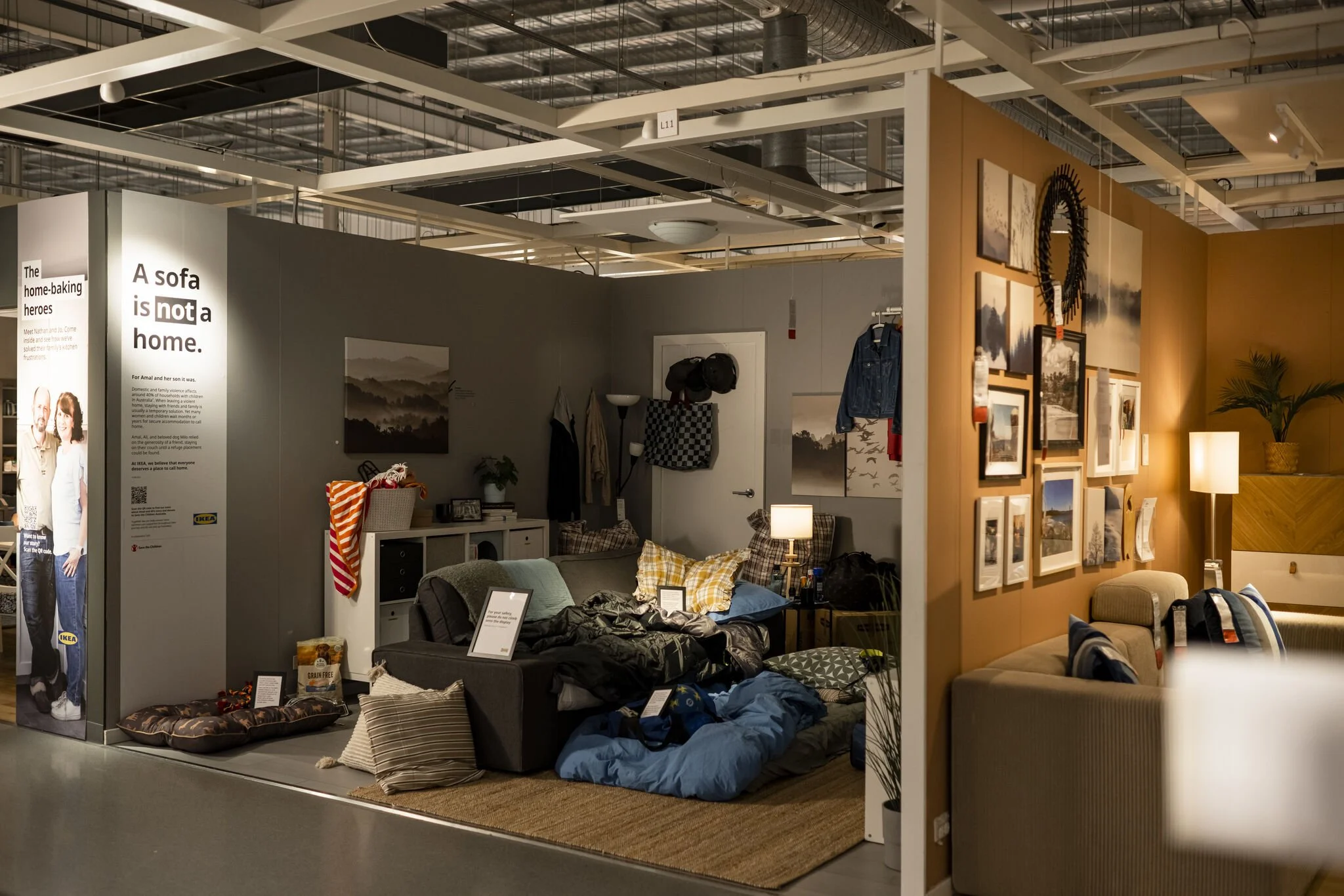 IKEA showroom display featuring a cluttered living room with sofas, bedding, pillows, wall art, lamps, and home decor