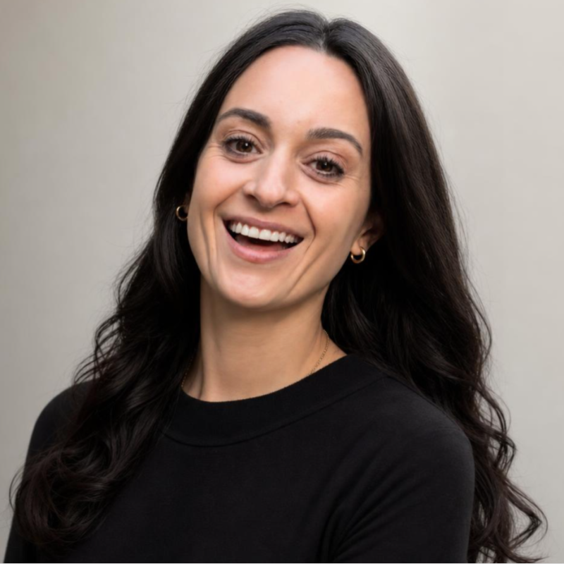 Smiling woman with long dark hair wearing a black top and gold hoop earrings against a gray background