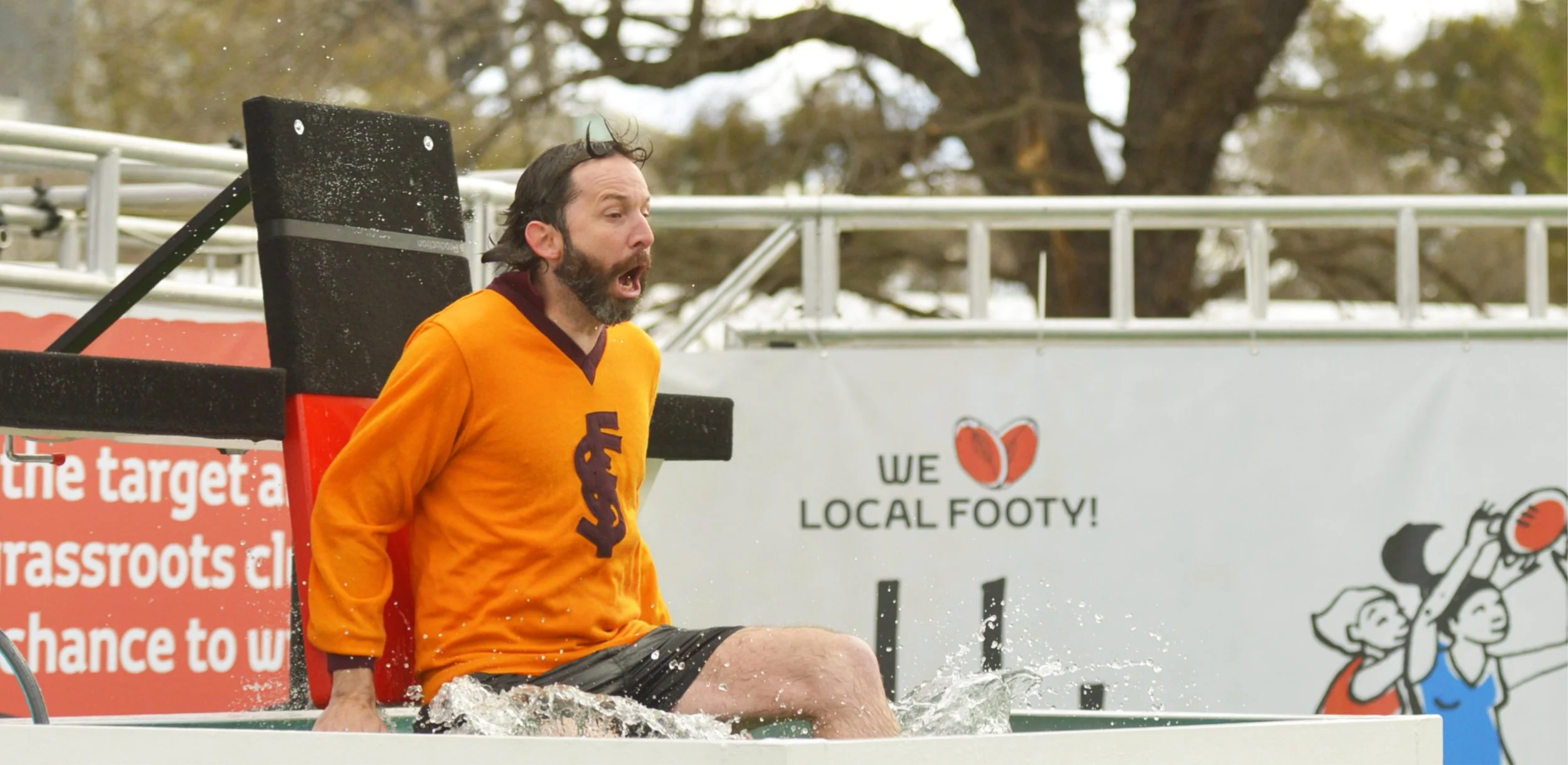 Bearded man in an orange soccer jersey sits in a water-filled tub, splashing at an outdoor sports event.