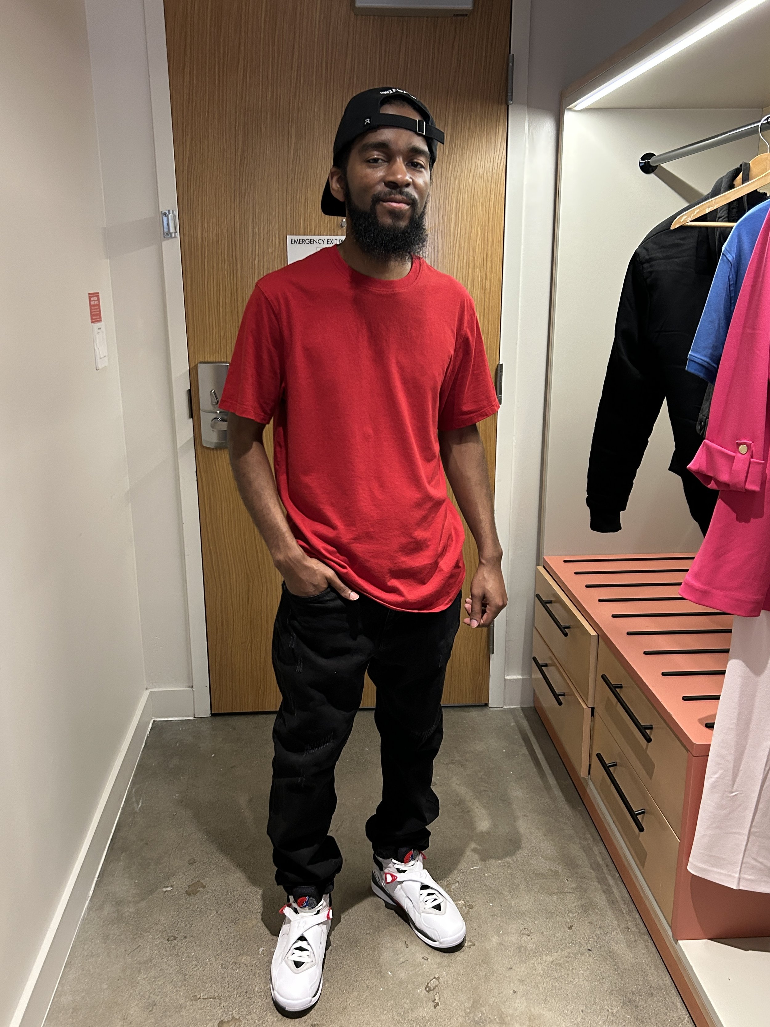 John Rollins. A young man with a beard, wearing a black baseball cap backwards, a red T-shirt, black jeans, and white sneakers, standing indoors in front of a wooden door and a clothing rack with jackets and shirts.