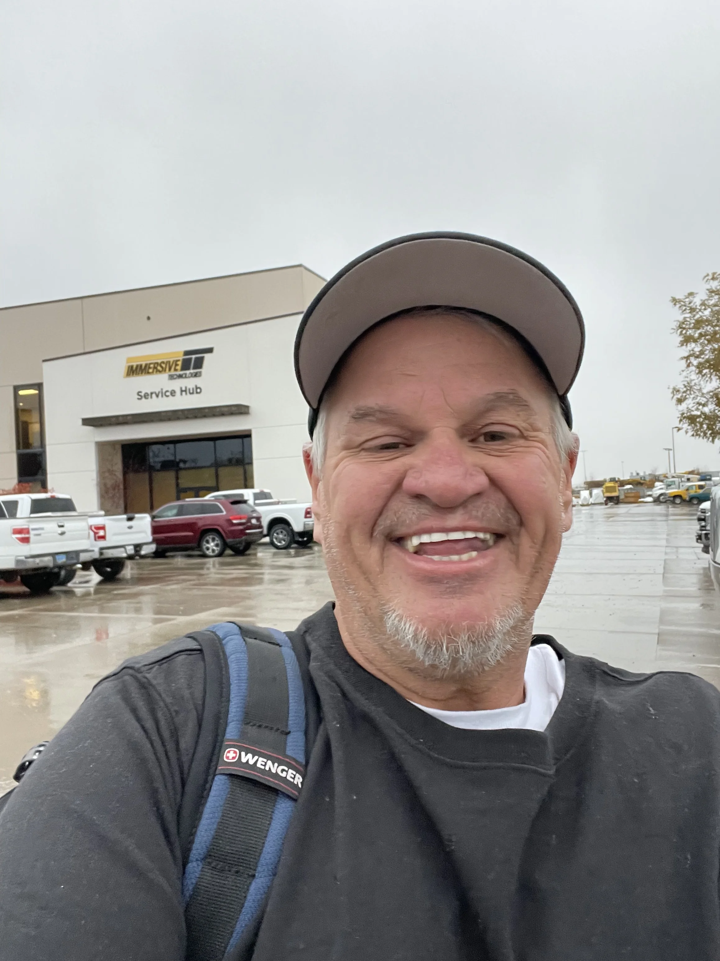 A smiling man in a black jacket and gray baseball cap taking a selfie outside in a parking lot on a rainy day, with cars and an 'Immersive Tech Hub Service Center' building in the background.