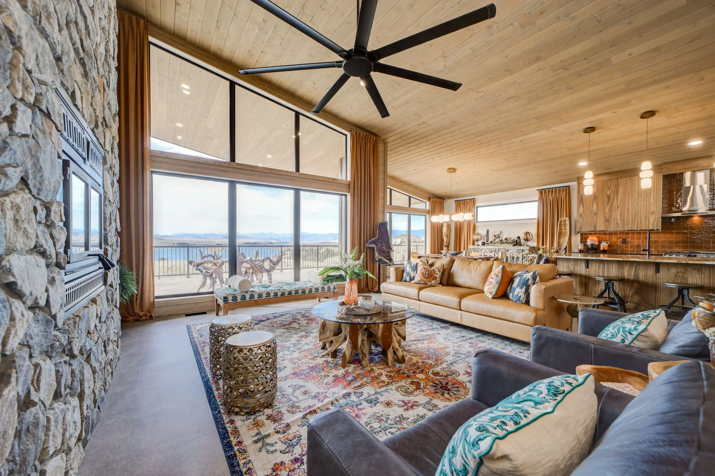 Spacious living room with large windows showing a scenic view, high wooden ceiling, stone wall, modern ceiling fan, and eclectic furniture including a leather sofa with colorful pillows, a patterned area rug, and a rustic wooden coffee table.