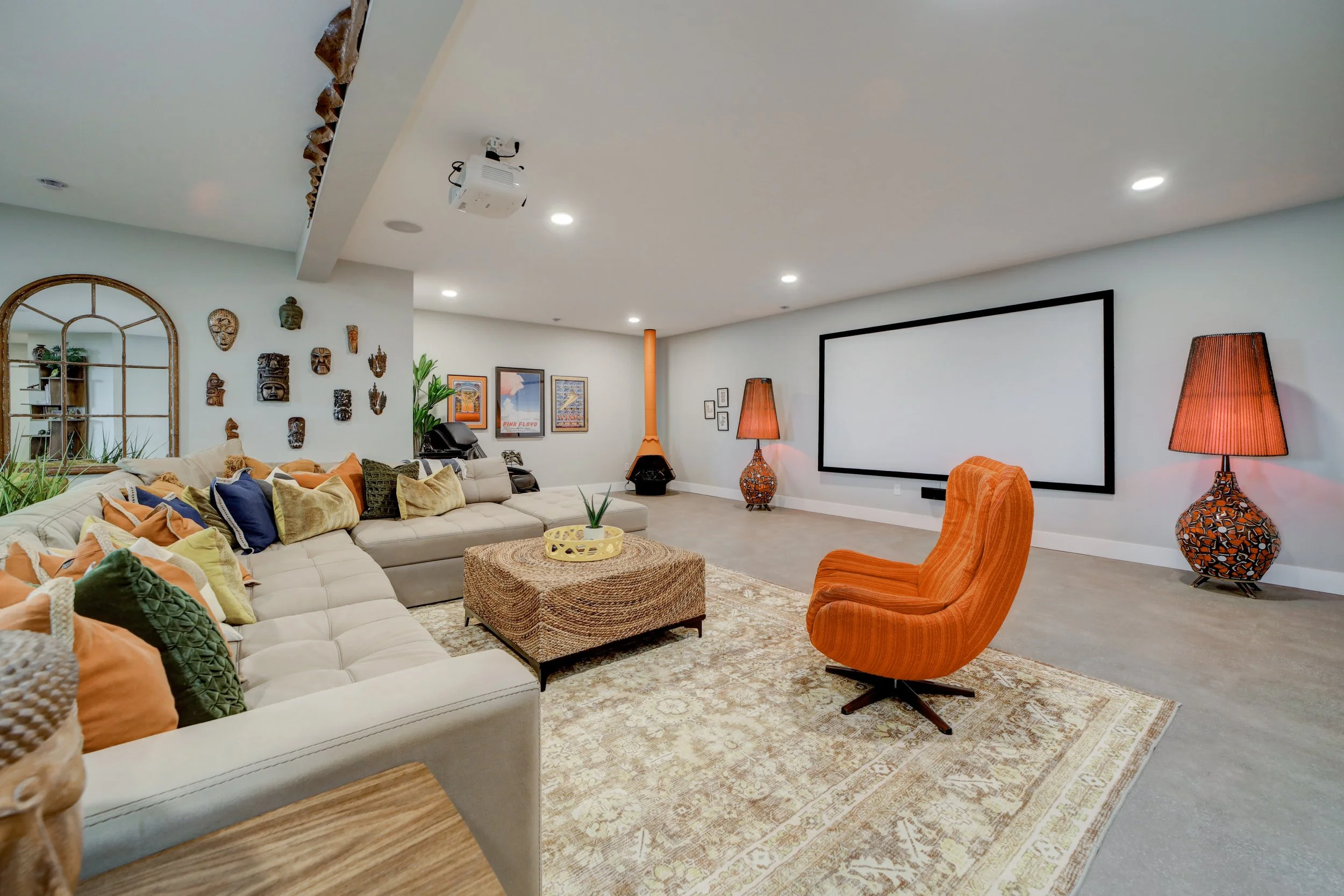 A spacious living room decorated with a large white sectional sofa with multiple colorful pillows, two orange upholstered chairs, a woven coffee table, and a patterned rug. The walls feature framed artwork and emotional masks, a large mirror, and a fireplace with an orange chimney. There are two tall lamps with orange bases and a projector mounted on the ceiling.