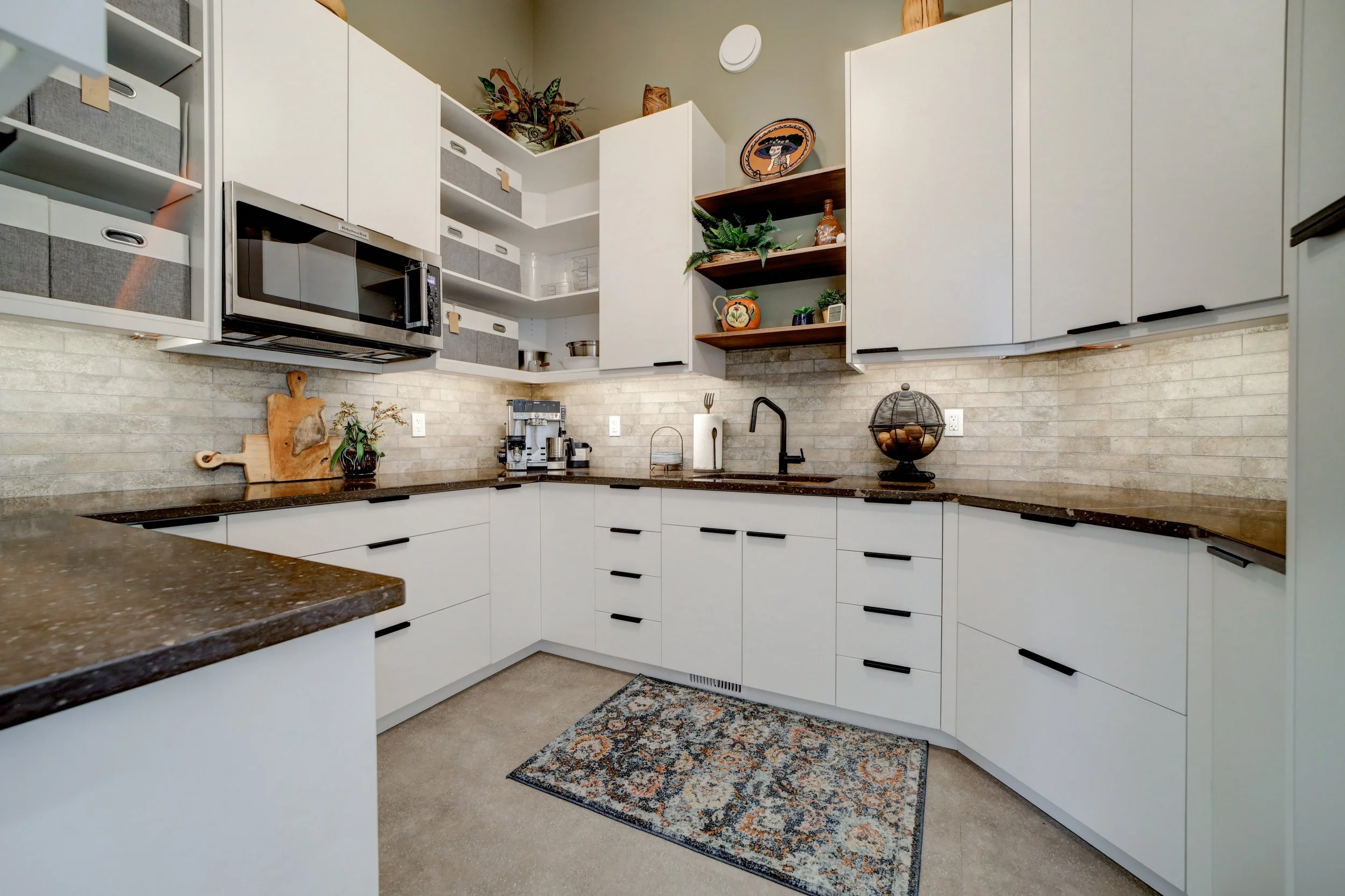 A kitchen with white cabinets, black handles, a brown speckled countertop, and beige brick backsplash. There is a black sink with a statement faucet, a small coffee station, and decorative items including plants, pottery, and artwork. An open shelf holds more plants and decorative objects, with ceiling-mounted cabinets above.