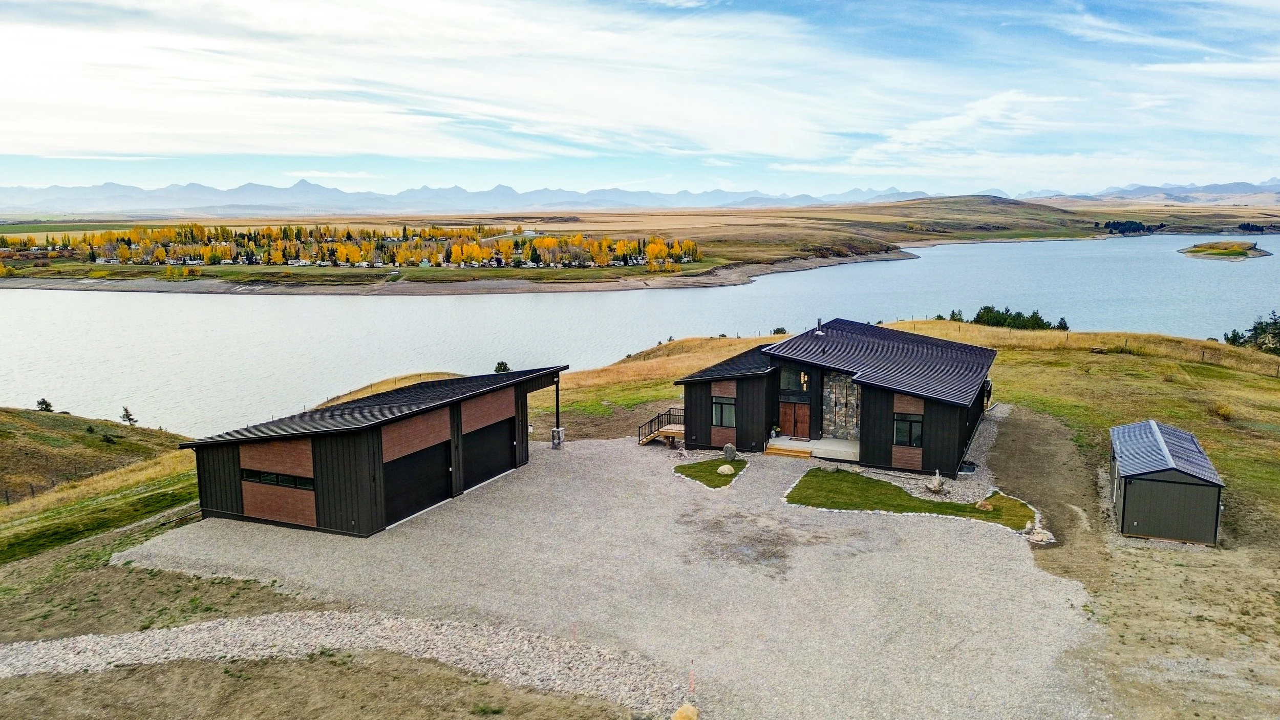 Modern black houses with brown accents situated on a hill overlooking a large body of water with a landscape of trees, fields, and mountains in the background under a partly cloudy sky.