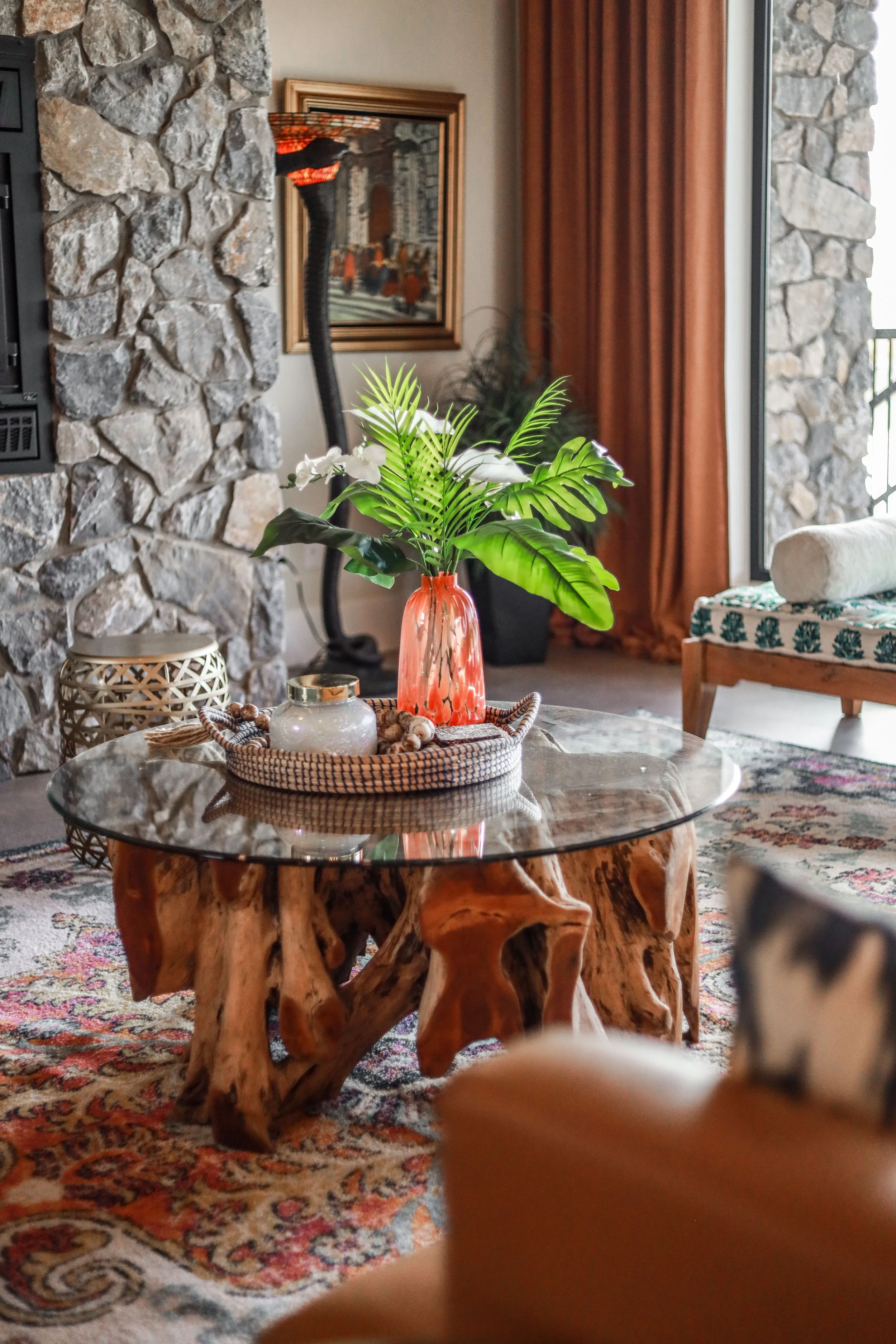 Living room with a stone fireplace, hardwood floors, a patterned rug, a wooden coffee table with a glass top, a decorative pumpkin, plants, curtains, a framed mirror, and a sofa.