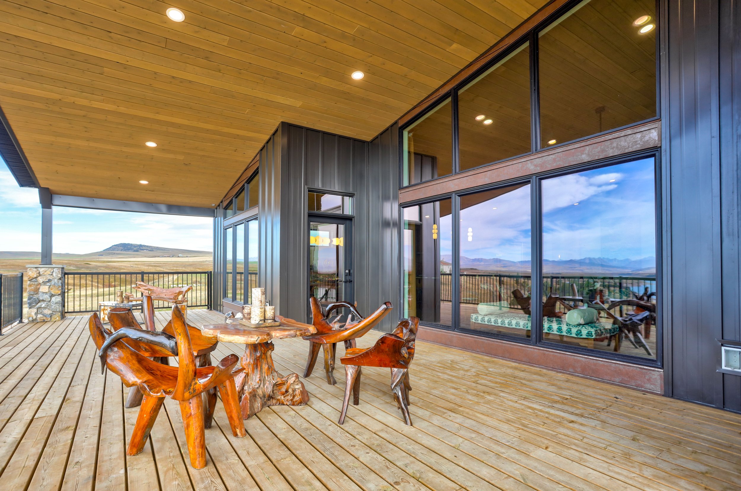 A spacious deck with a wooden table and chairs made from carved wood, overlooking a mountain landscape. The deck has a wooden ceiling with recessed lighting, glass doors, and windows reflecting the sky and landscape.