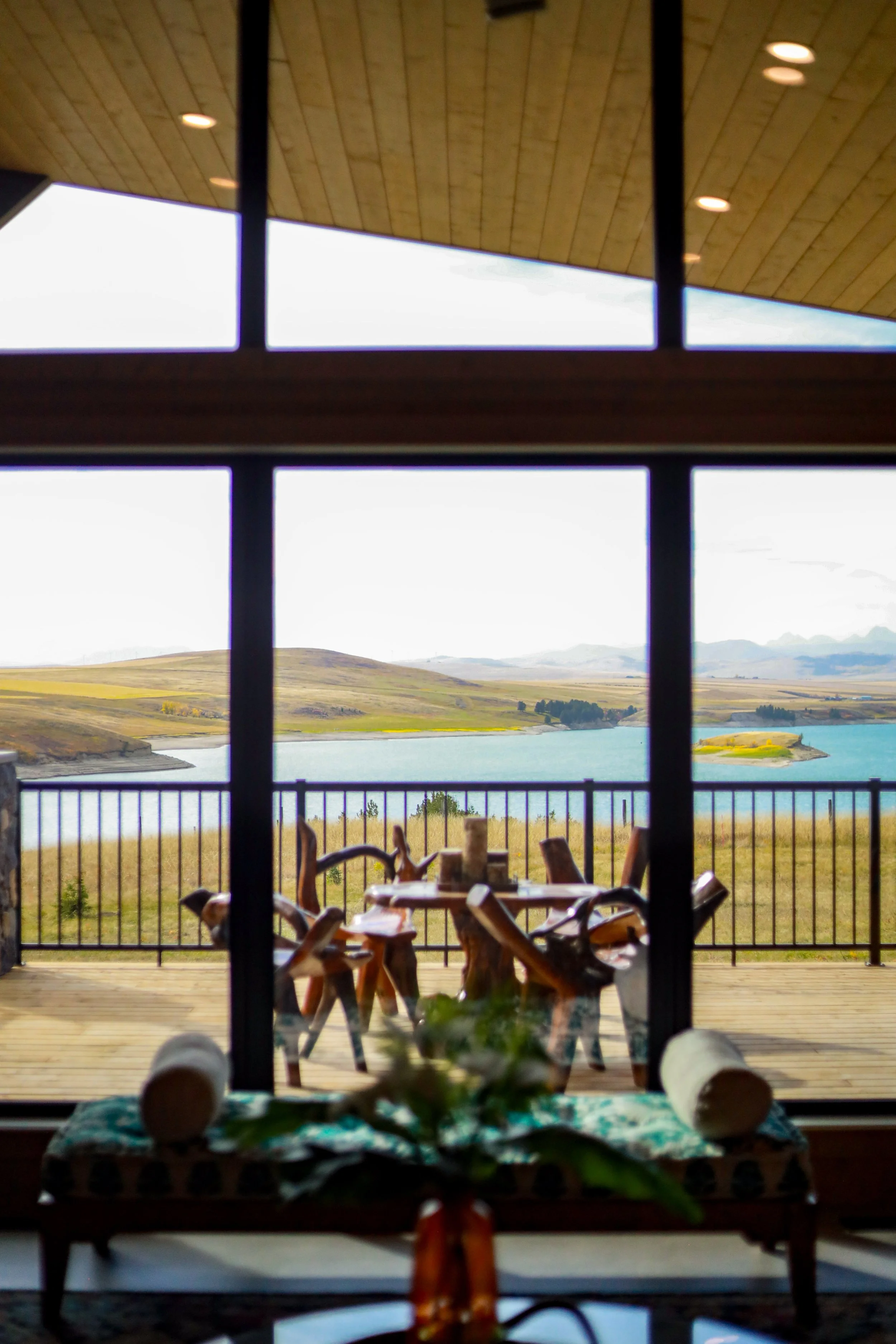 View of rolling hills and a body of water seen through large glass windows from inside a wooden room. There are wooden chairs outside on a deck, enclosed by a metal railing, with a wooden ceiling inside.