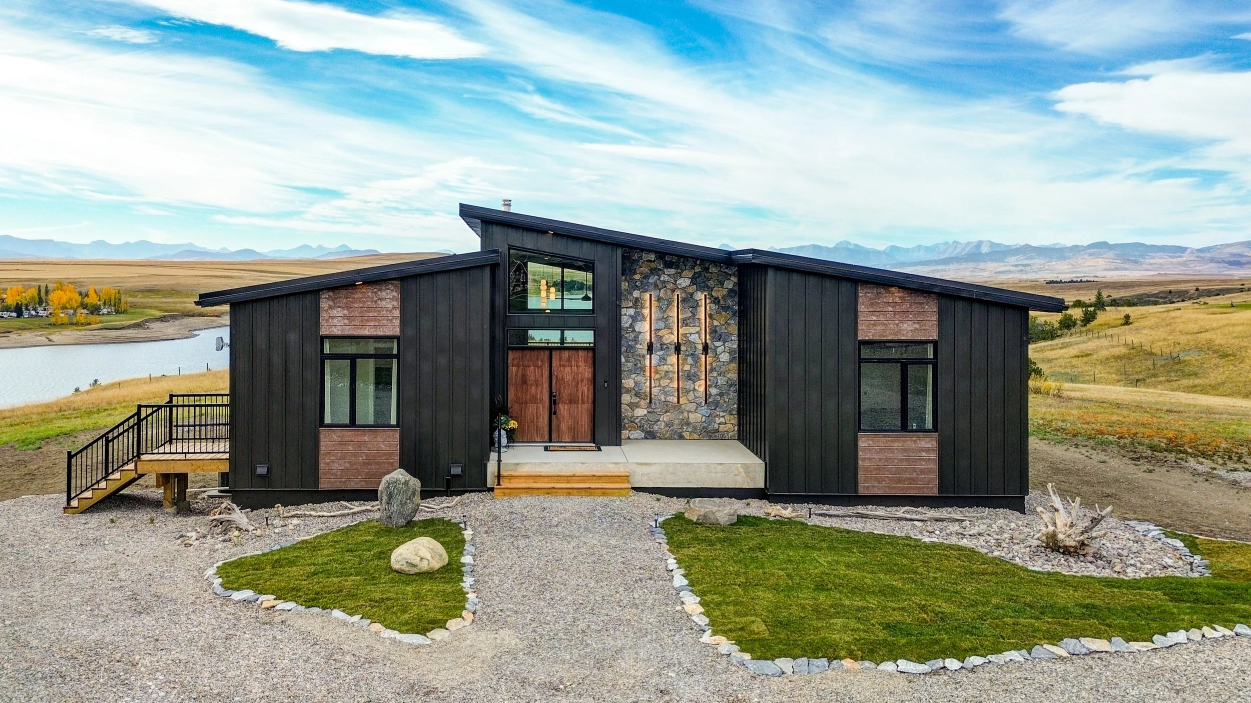 Modern black house with stone details and large windows, surrounded by a landscaped yard with gravel paths and small grass patches, overlooking a lake and mountains in the distance under a partly cloudy sky.