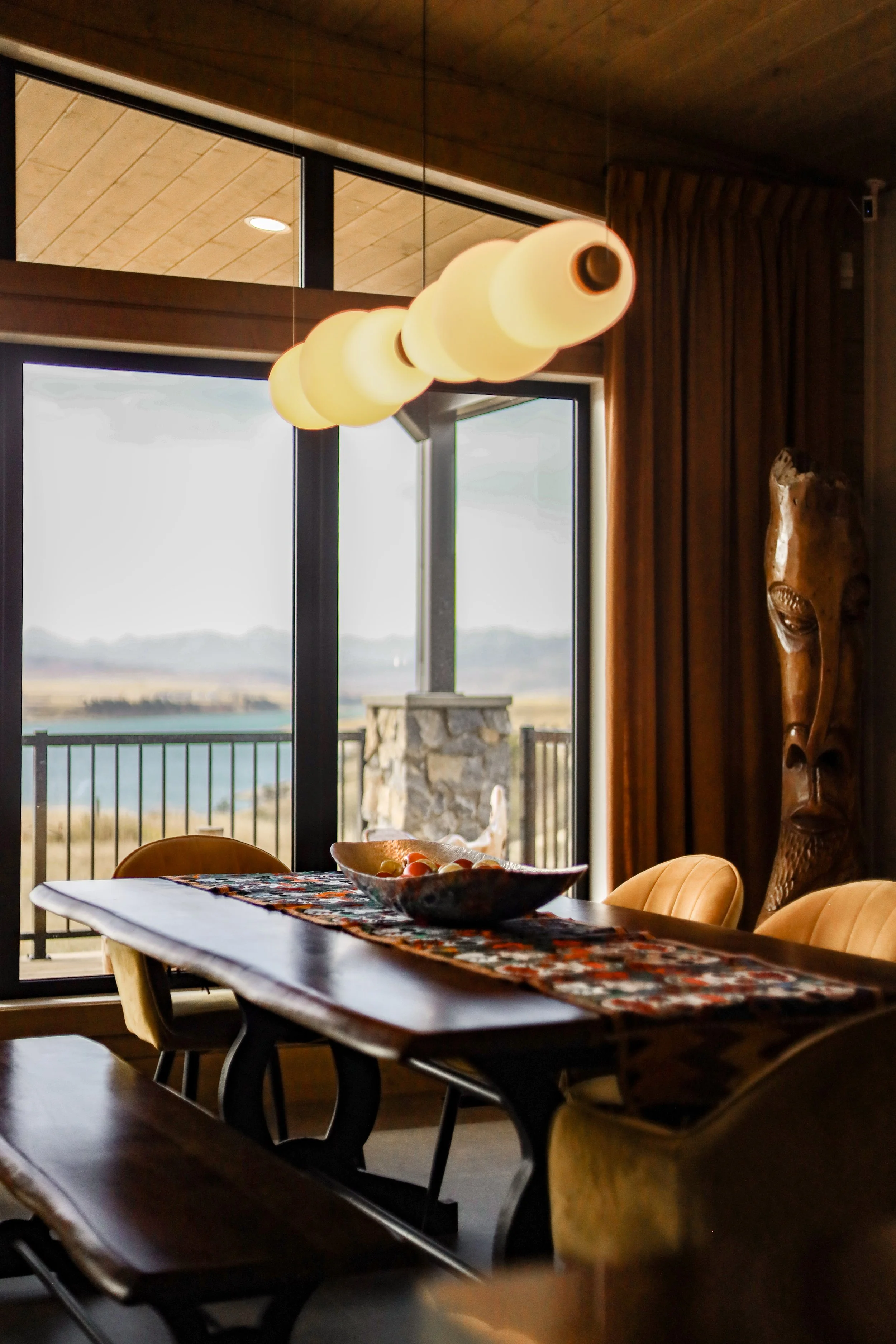 A dining room with a wooden table, beige chairs, and a colorful table runner. A bowl of fruit is on the table. Large windows reveal an outdoor patio and a scenic landscape with mountains in the background. A decorative wooden sculpture is on the wall.