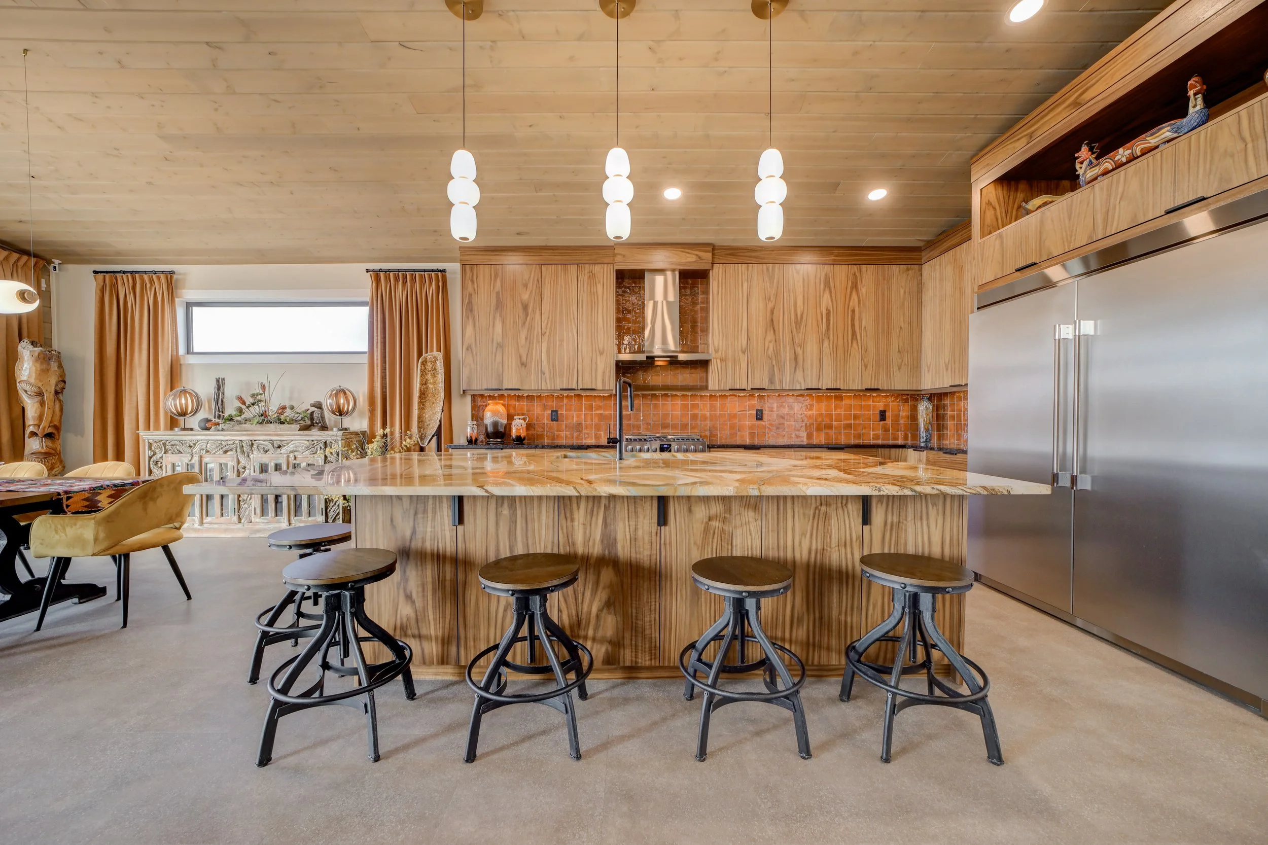 Modern kitchen with wooden cabinets and a large granite island large enough for six, and pendant lights hanging from an wood-cladded ceiling, stainless steel appliances.