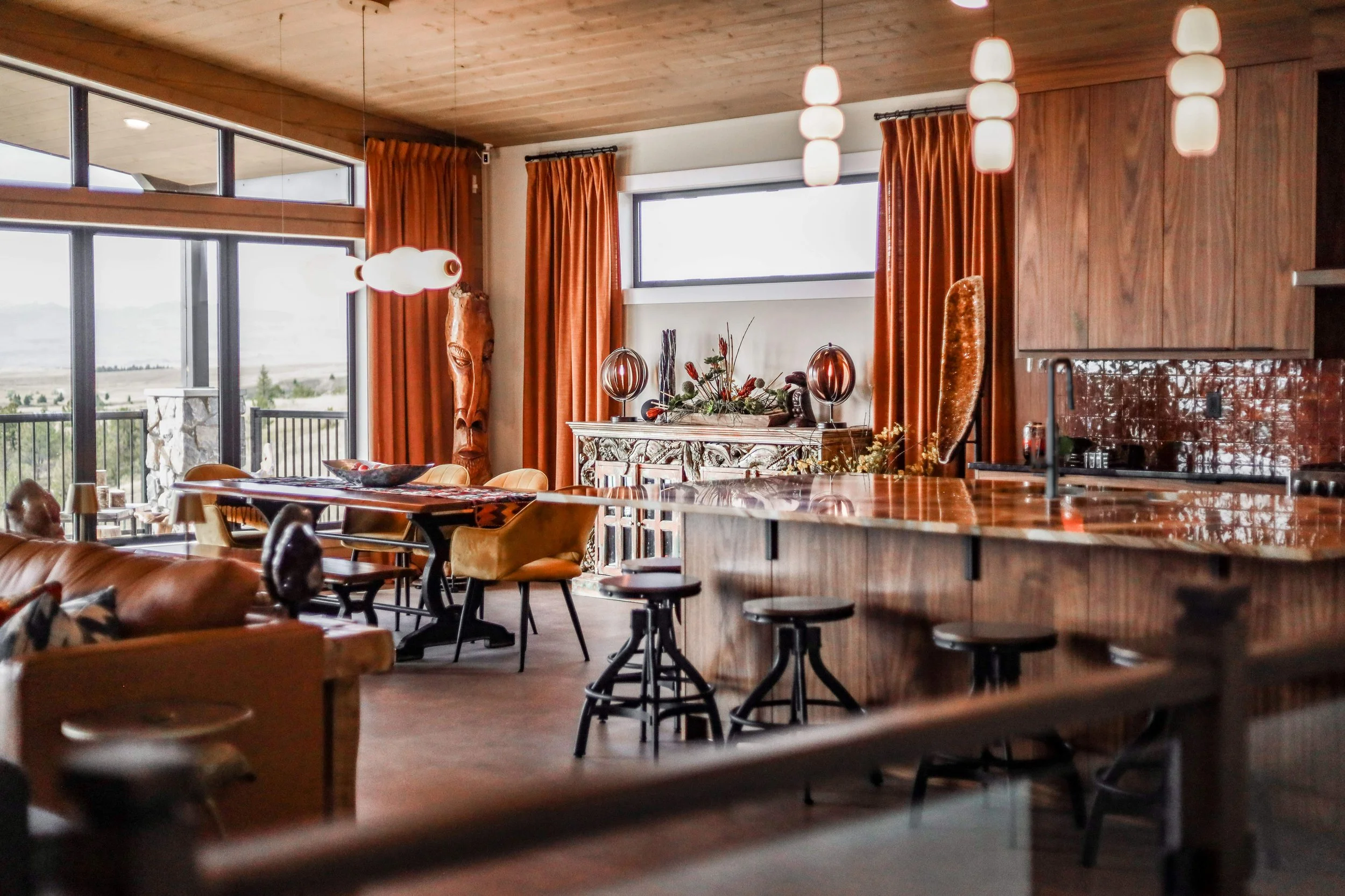 Interior of a modern home with a wooden ceiling, large windows, orange curtains, a decorative sideboard with vases and floral arrangements, a kitchen island with black stools, and hanging pendant lights, with a view of an outdoor landscape.
