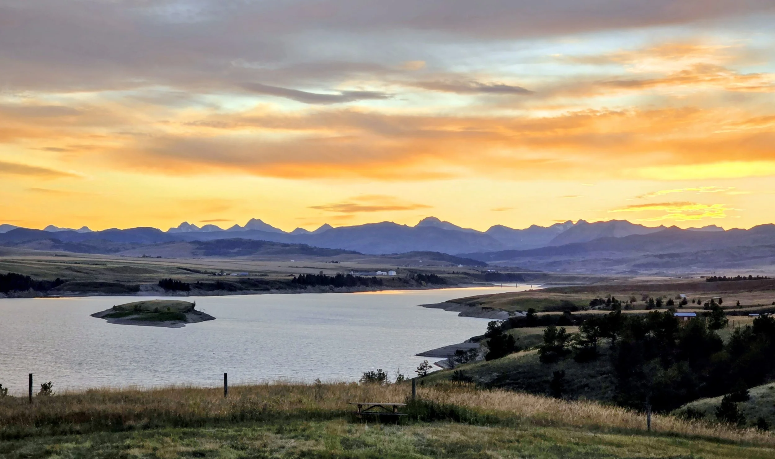 A scenic landscape at sunset with a river in the foreground, rolling hills, and a mountain range in the distance under a colorful sky with orange, yellow, and blue hues.