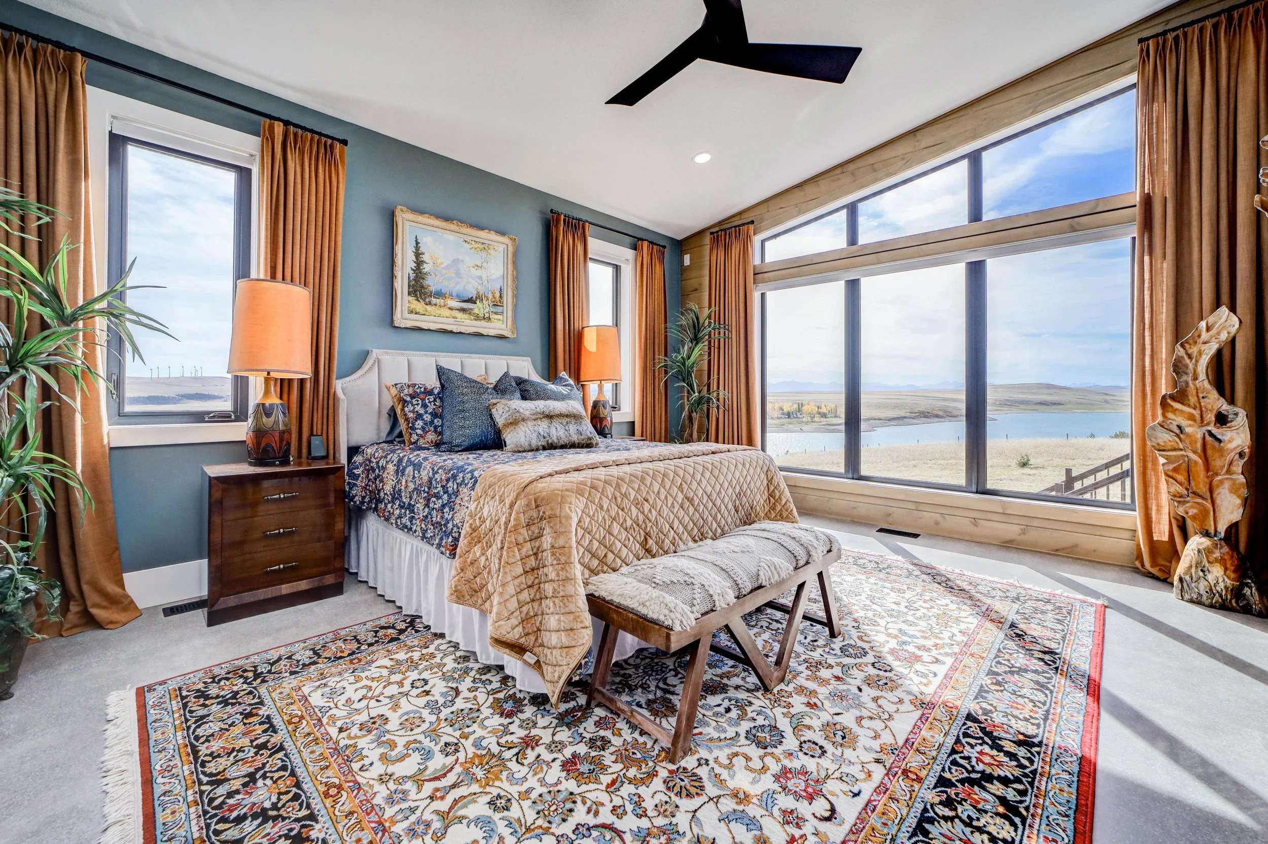 A bedroom with large windows showing a view of water and landscape, decorated with colorful rugs, a bed with patterned bedding, orange curtains, and plants.