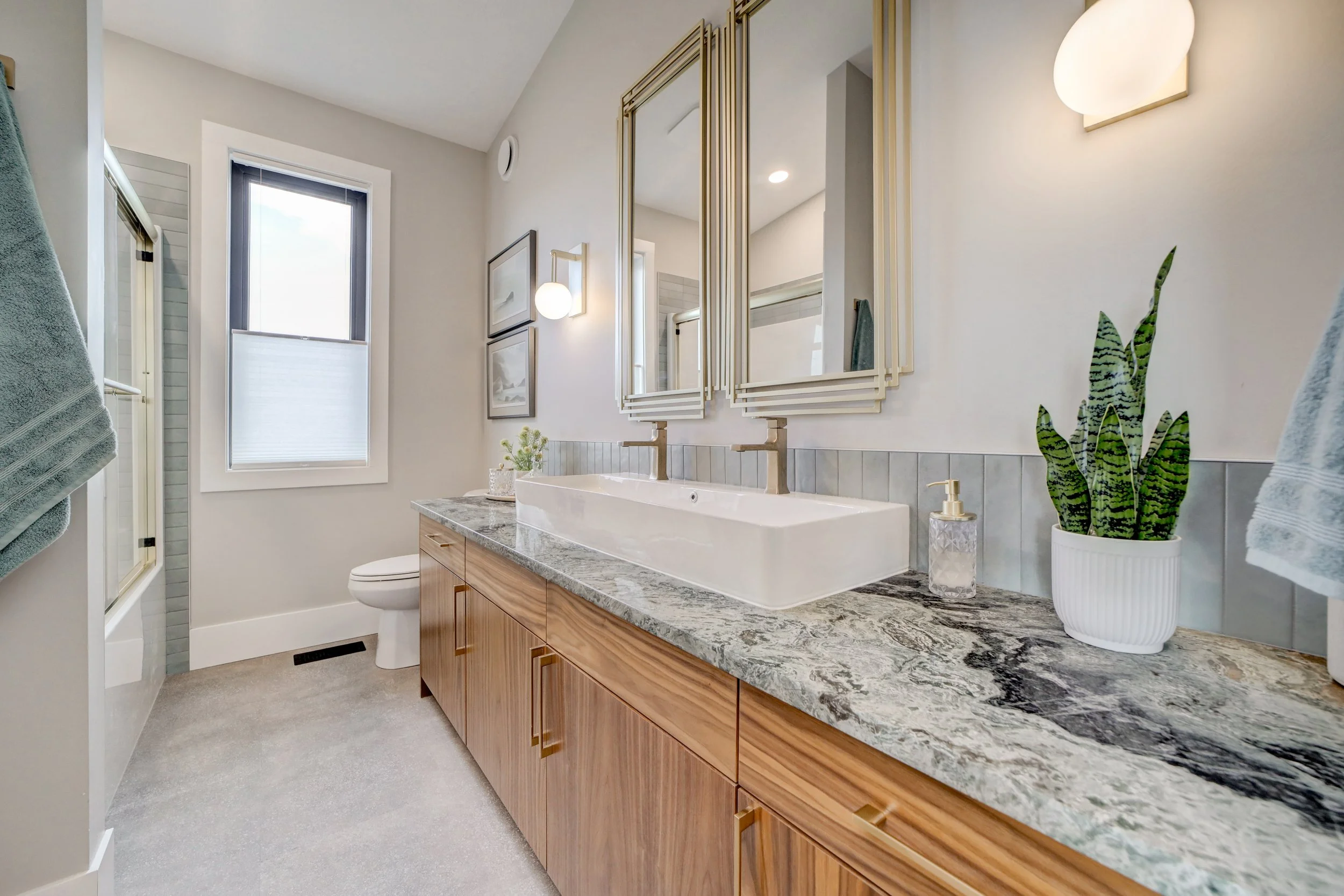 A modern bathroom with a marble countertop vanity, a large mirror, a potted plant, and decorative wall art. There are two windows with shades, a nearby toilet, and soft lighting fixtures.