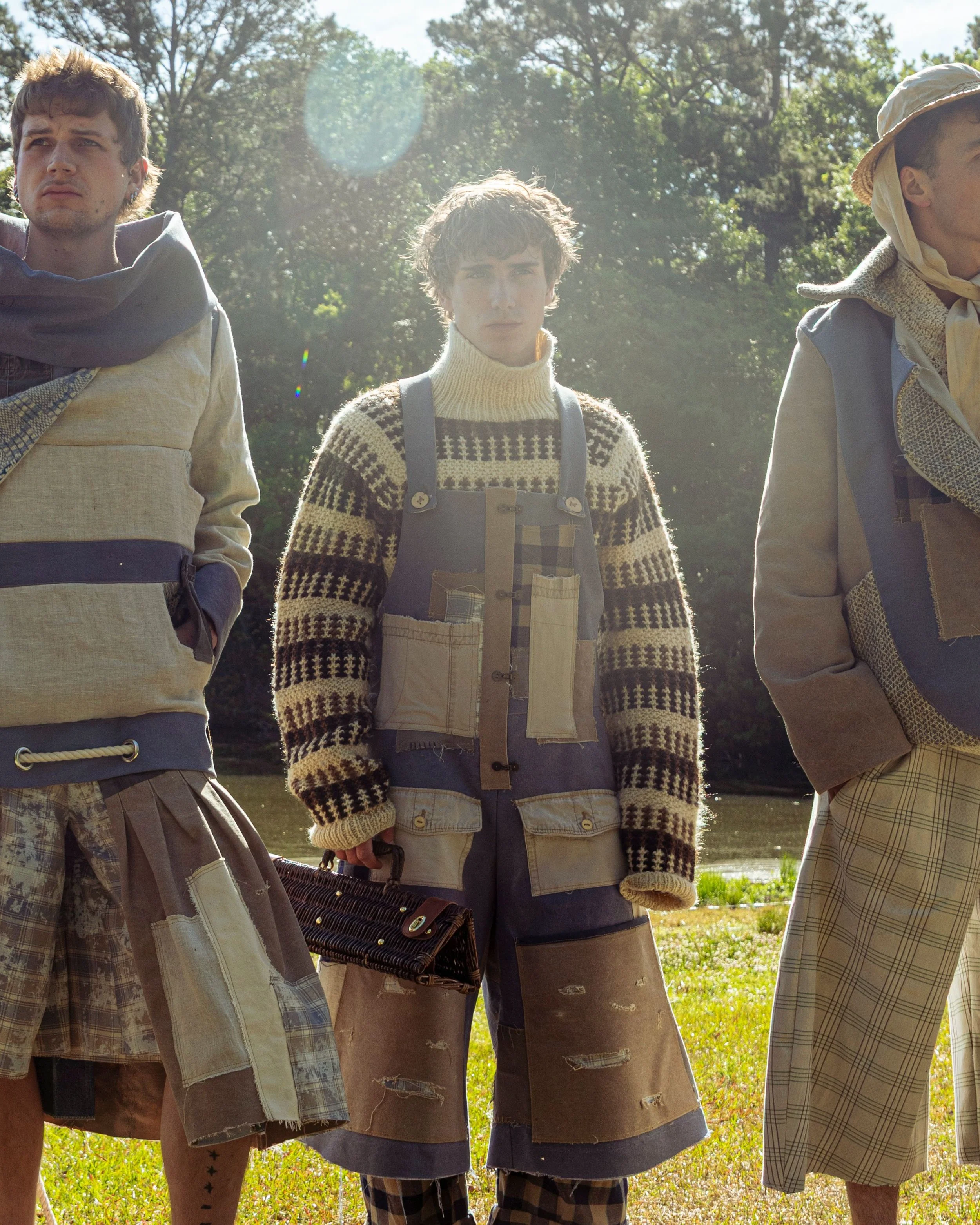 Three young men standing outdoors near a body of water with trees in the background, dressed in casual, vintage-style clothing, with sunlight illuminating the scene.
