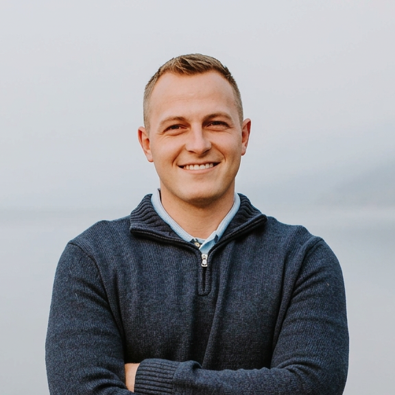 Young man smiling outdoors with arms crossed, wearing a navy zip-up sweater and a collared shirt, against a cloudy sky background.