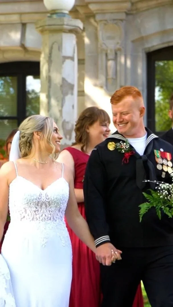 A bride and groom holding hands and smiling during their wedding ceremony outdoors, with a woman in a red dress in the background.