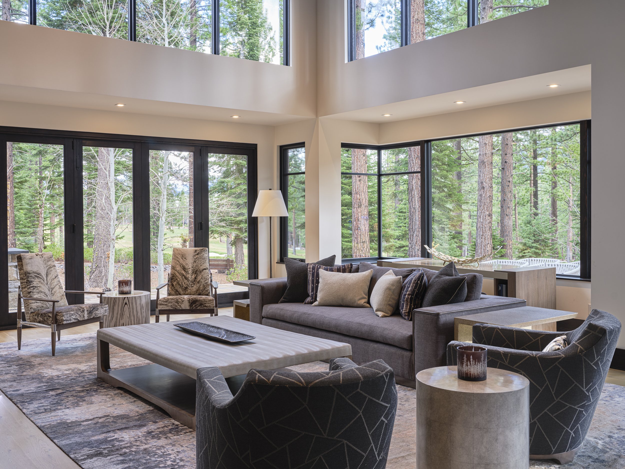 Interior of a living room with large windows showing a forest of tall trees outside, including a sectional sofa with pillows, a coffee table, armchairs, and a standing lamp, all in a modern design.