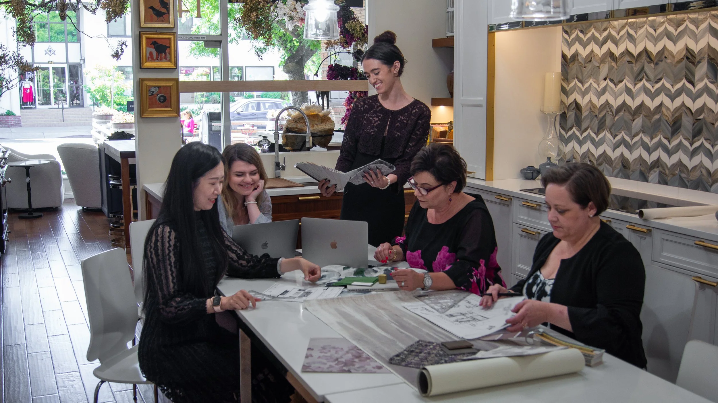 A group of five women gathered around a table in a well-lit, modern cafe or studio. One woman is standing, smiling and holding a clipboard, while the others are sitting and working on laptops and papers. The setting features decorative wall art, large windows, and a colorful, patterned backsplash.