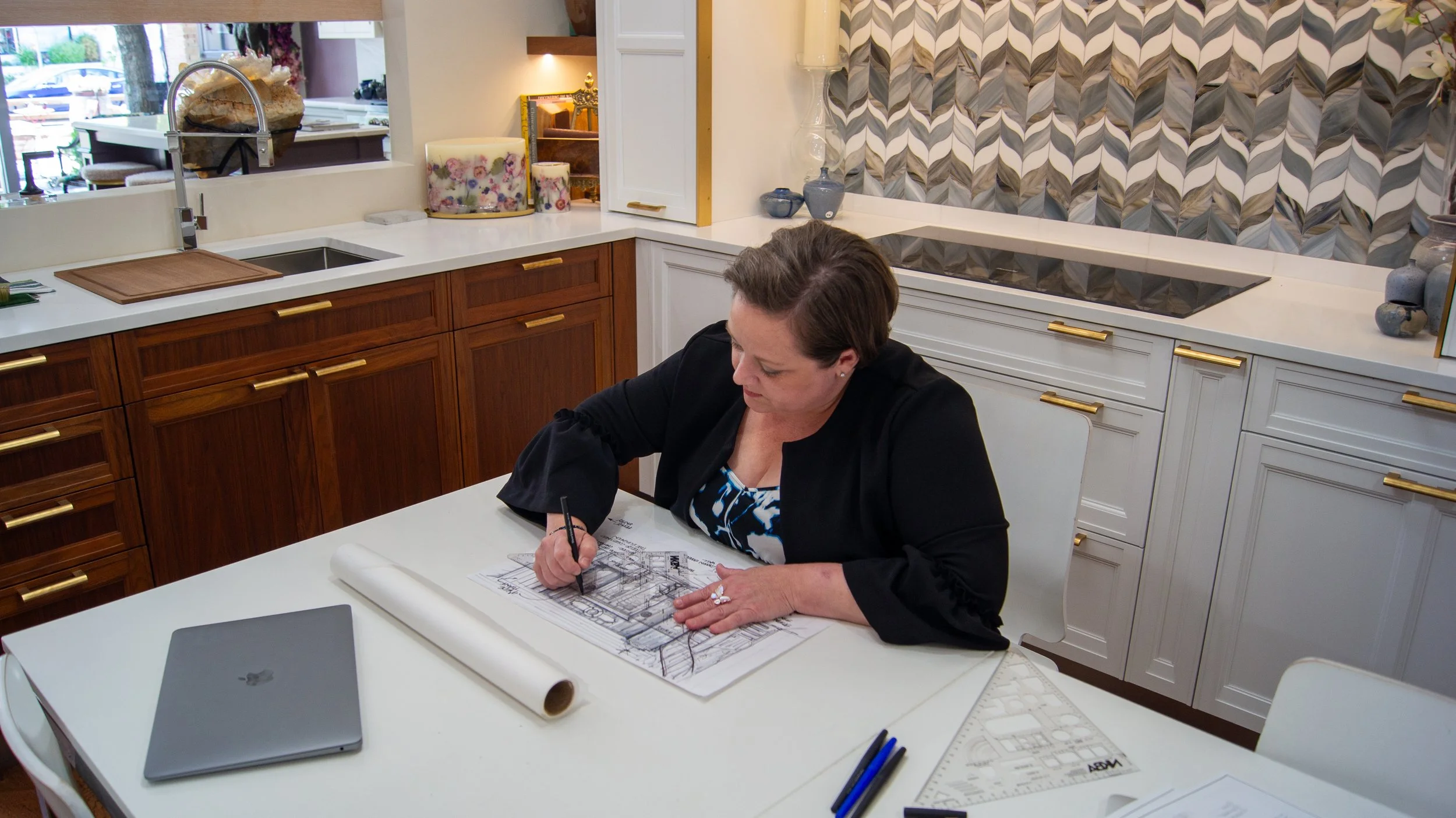 A woman in a black blazer and printed blouse sitting at a white table, working on architectural plans with a pencil. The kitchen background features wooden and white cabinets, decorative vases, and a patterned tile wall.