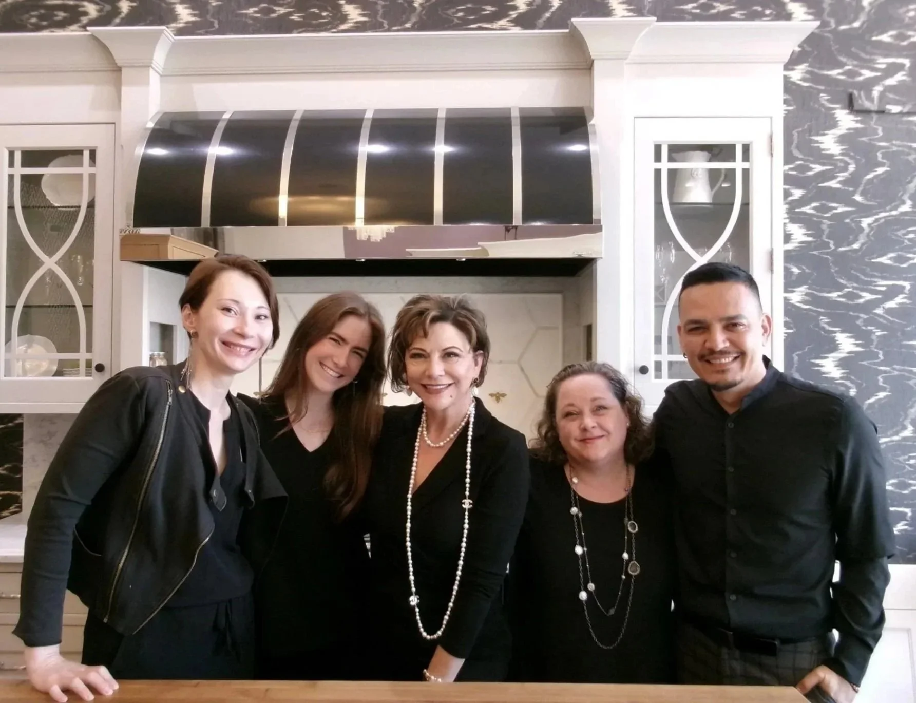 Five smiling people standing together indoors in front of a black and white marble-patterned wall, posing for a group photo.