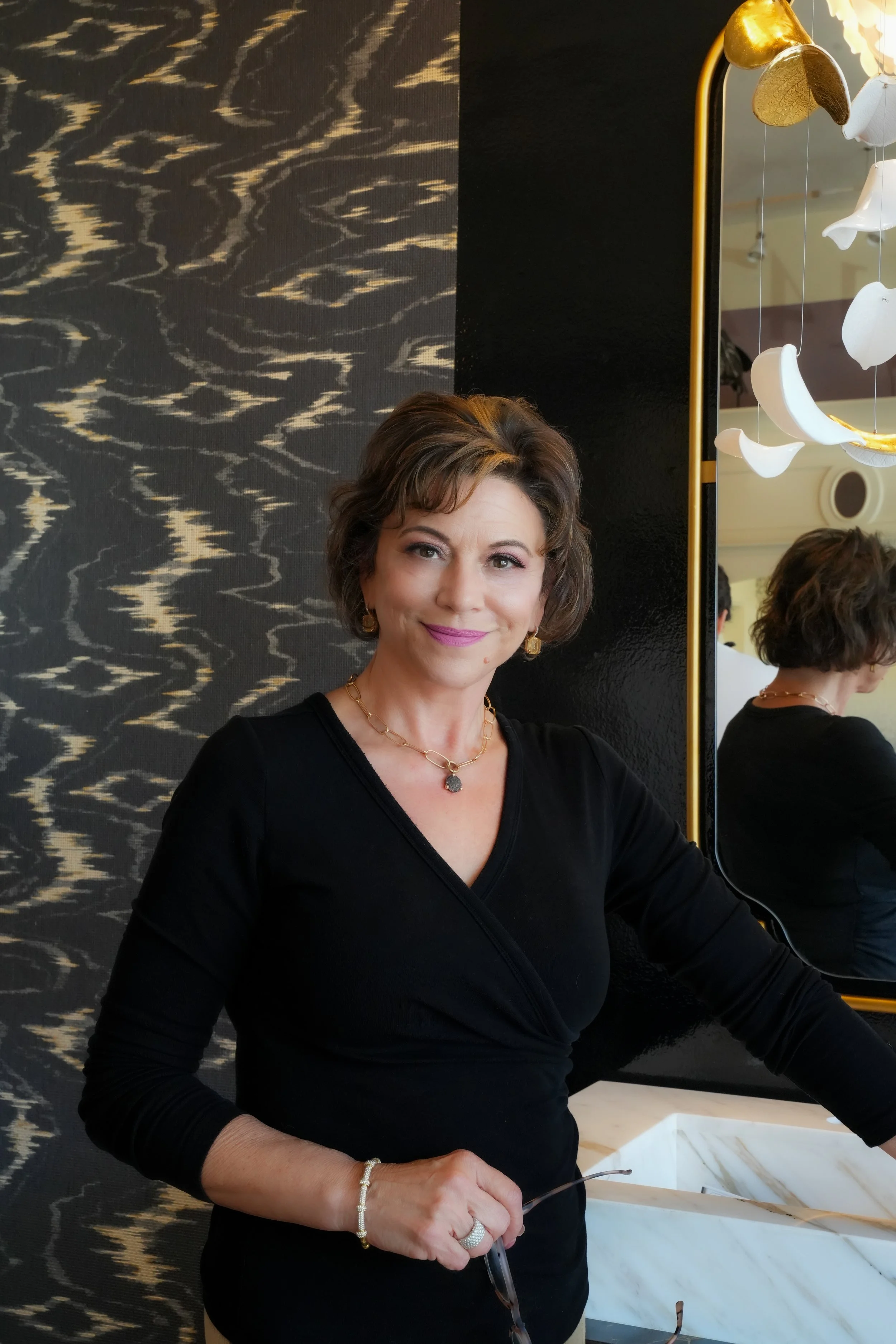 A woman with short brown hair, wearing a black top, gold jewelry, and pink lipstick, standing indoors near a black and gold wall, with a mirror reflecting her back and a hanging light fixture with gold and white elements.
