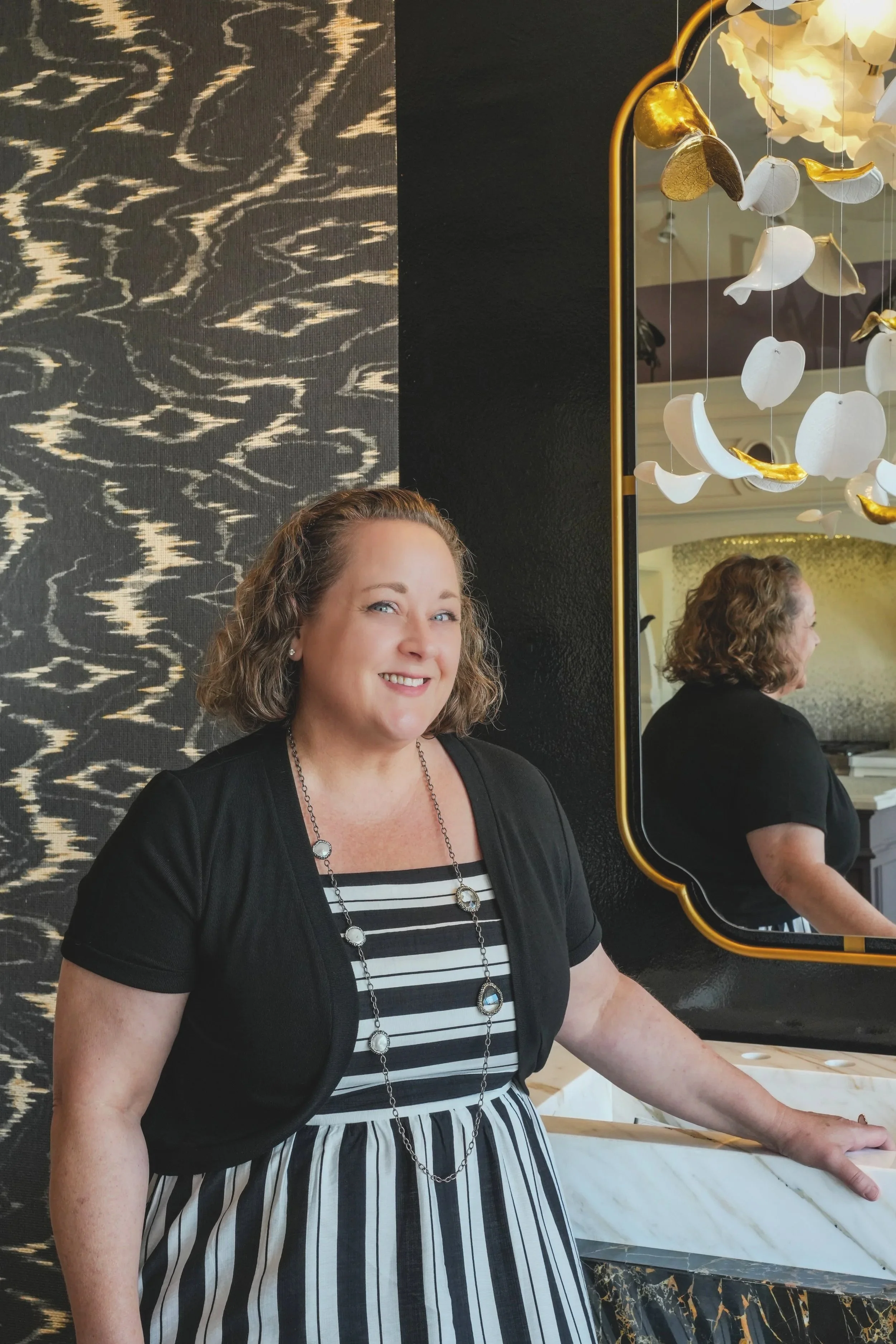 A smiling woman with curly hair and a navy blue top with a printed dress, is standing in front of a mirror. The mirror reflects her profile and a decorative hanging sculpture of white and gold flowers. The background features a textured black and gold wall and a marble surface.