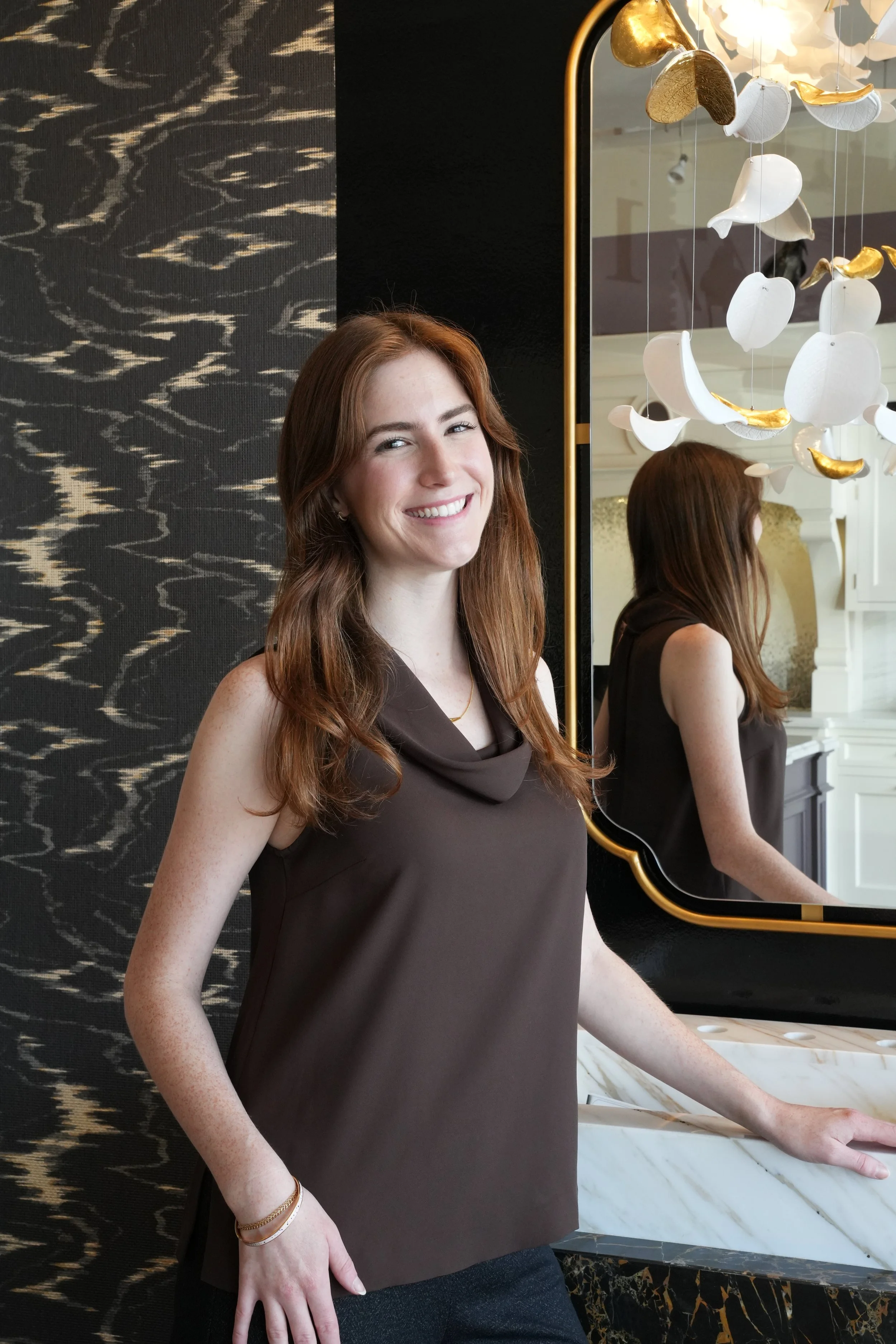 A woman with red hair wearing a brown sleeveless top smiling near a large mirror and a decorative chandelier, background featuring black-patterned wall and marble counter.