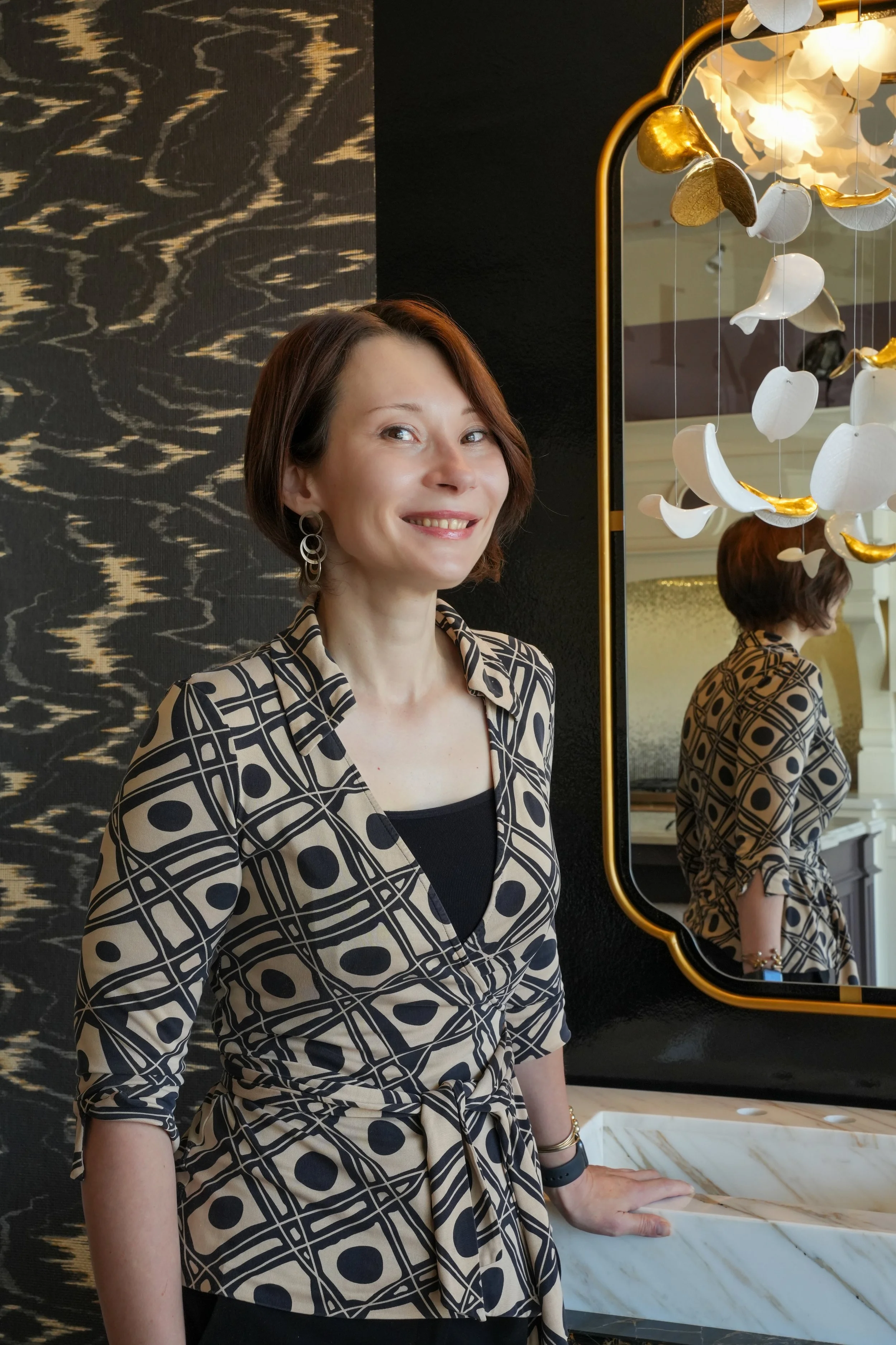 A smiling woman with short brown hair and wearing a patterned beige and black blouse, standing indoors near a mirror with hung decorative white and gold leaves and a black and gold wall background.
