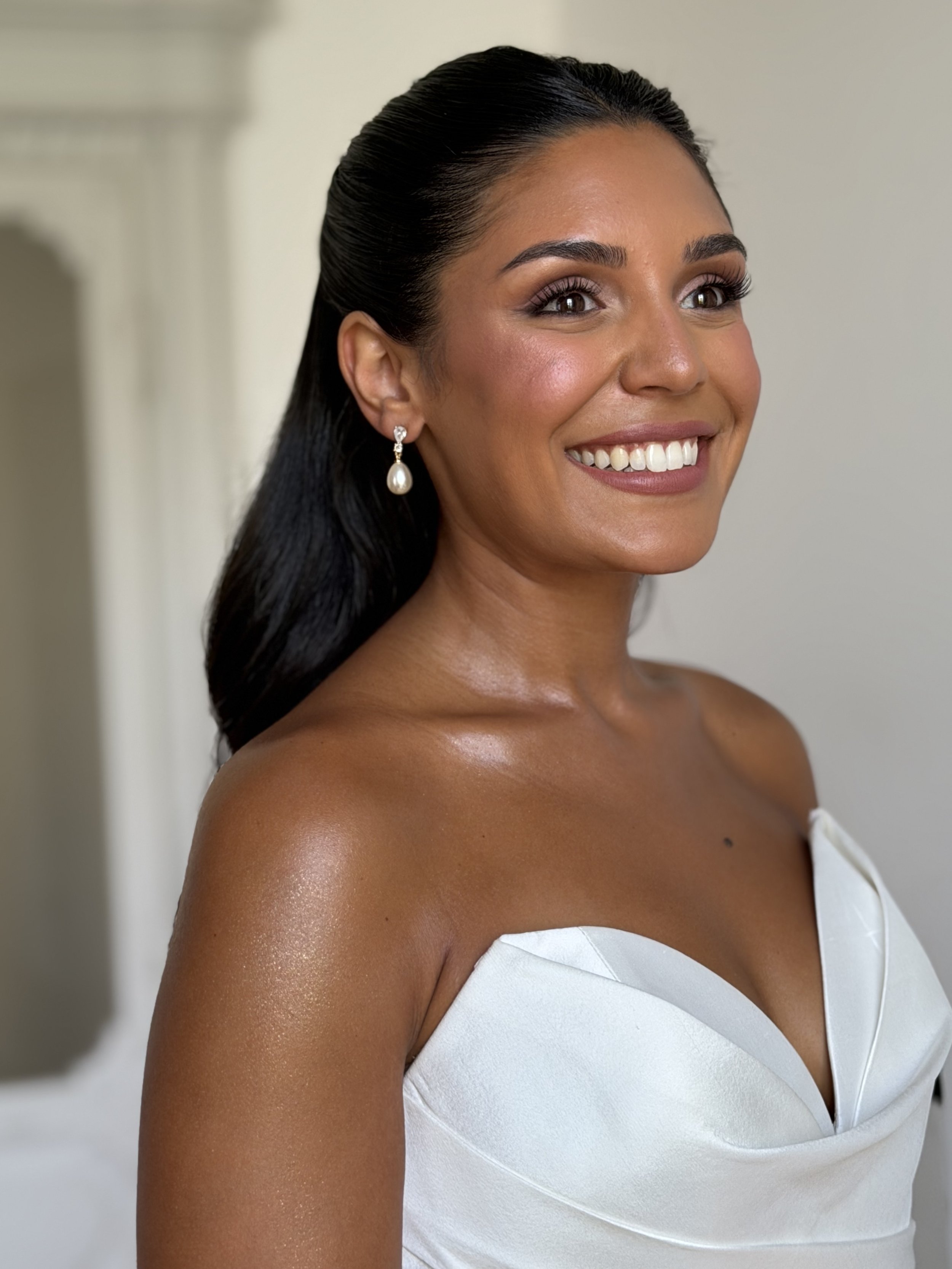Close-up of a woman with dark hair styled back, wearing pearl earrings, smiling, in a white dress, showing her shoulder and part of her chest.