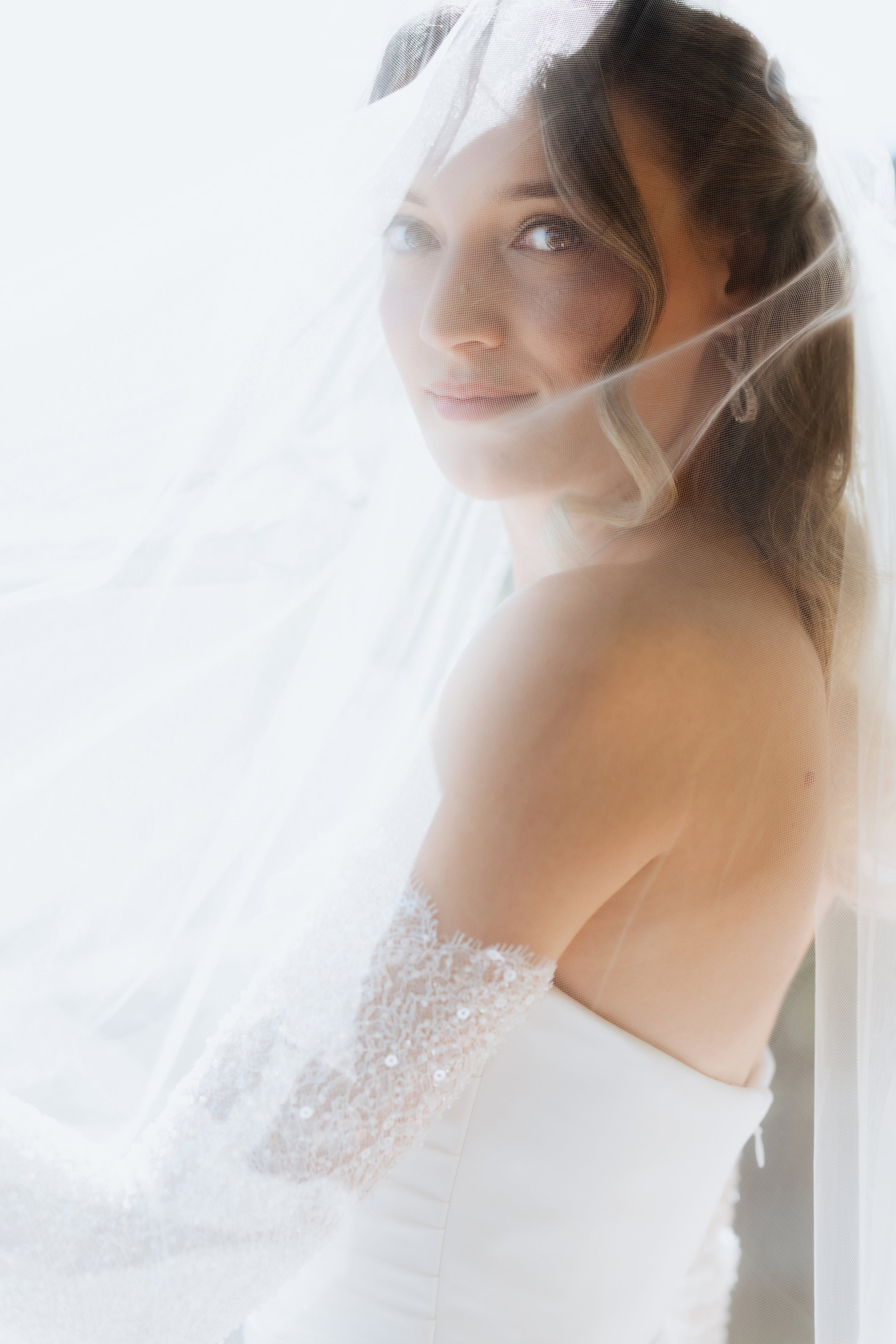 A woman with wavy brown hair and blue eyes, wearing white lace lingerie, draped in a sheer white veil with soft lighting.