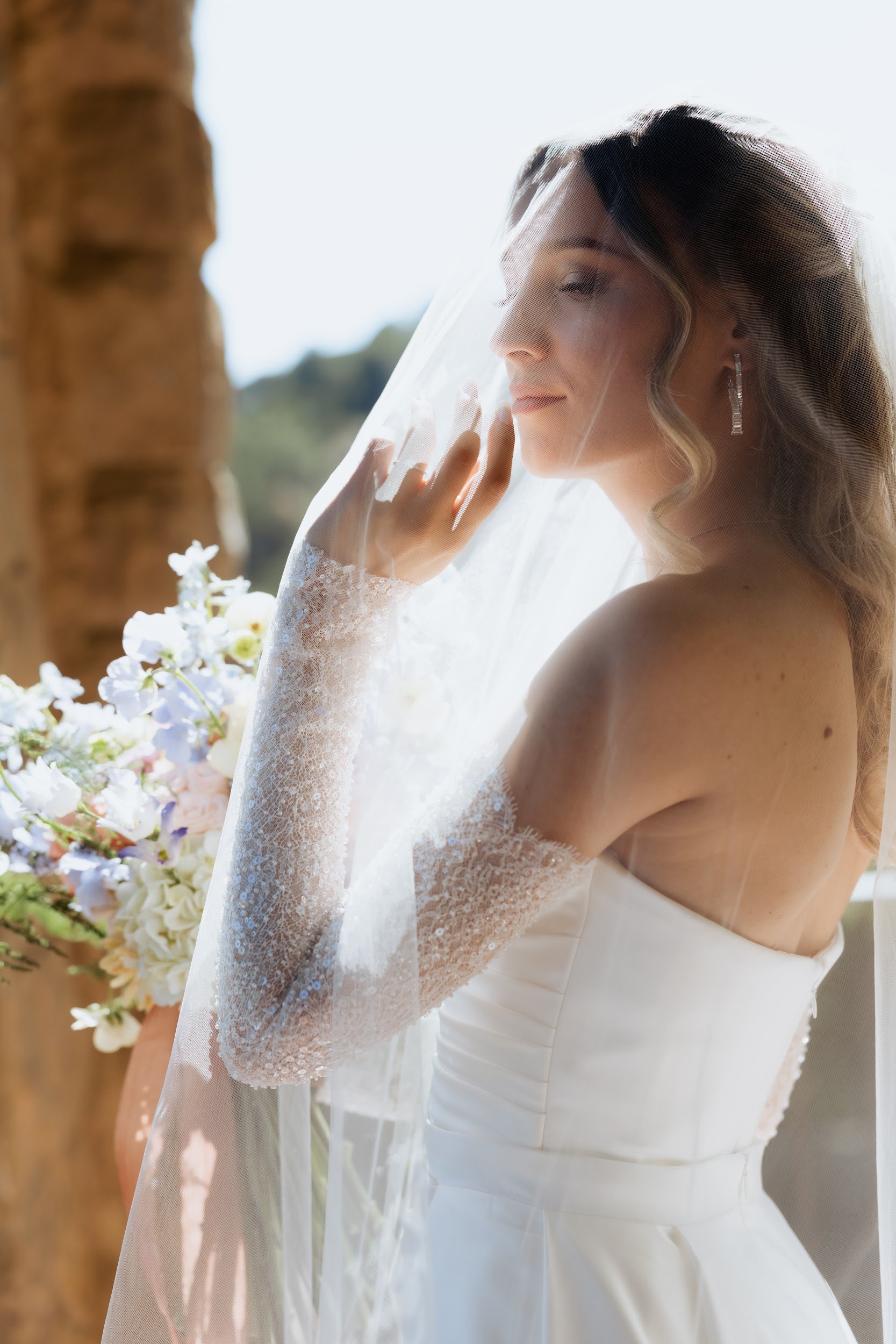 A bride in a white wedding gown with lace sleeves and a veil, standing outdoors with a bouquet of flowers, looking to the side near a wooden structure.
