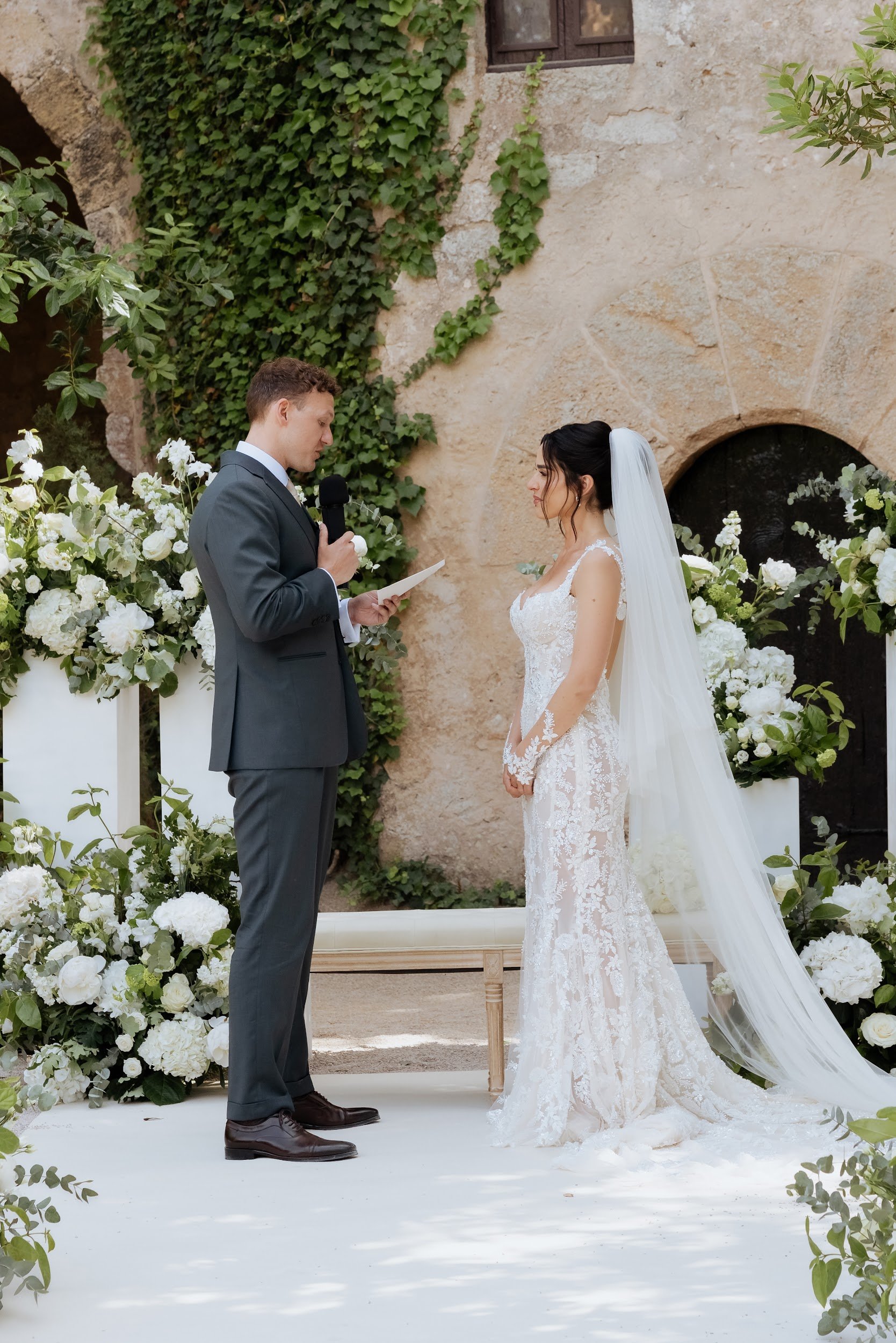 A groom in a dark gray suit and a bride in a white lace wedding gown and veil standing outdoors during a wedding ceremony, surrounded by white and green floral arrangements, with a stone wall and ivy in the background.