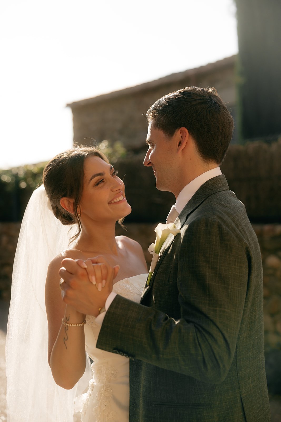 A bride and groom dancing outdoors at sunset, smiling and gazing into each other's eyes, with a stone building in the background.