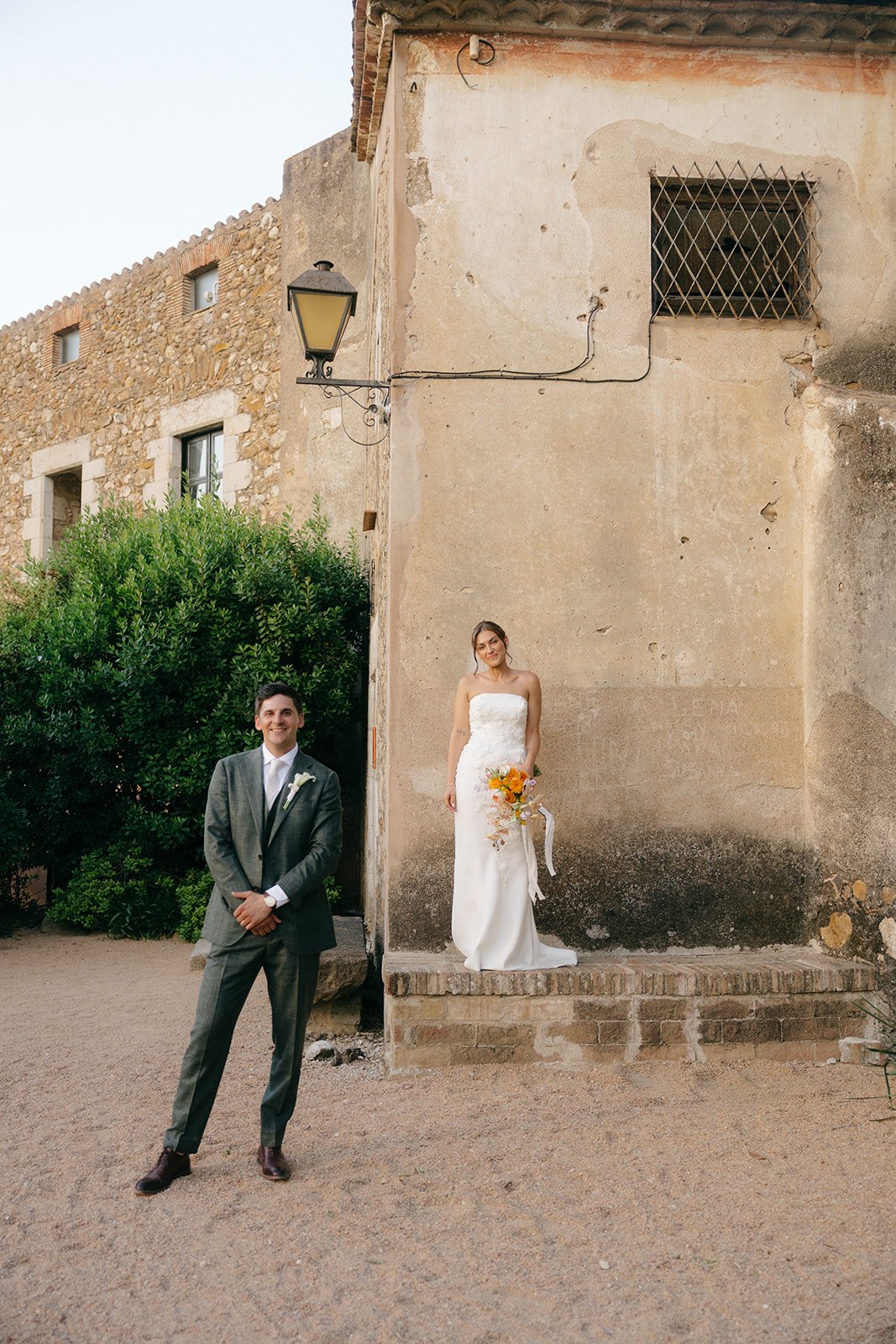A bride in a white strapless wedding gown holding a bouquet of yellow flowers, standing on a raised brick step against a weathered beige wall with a window and an outdoor lamp. A groom in a gray suit with a white shirt and boutonniere stands nearby o