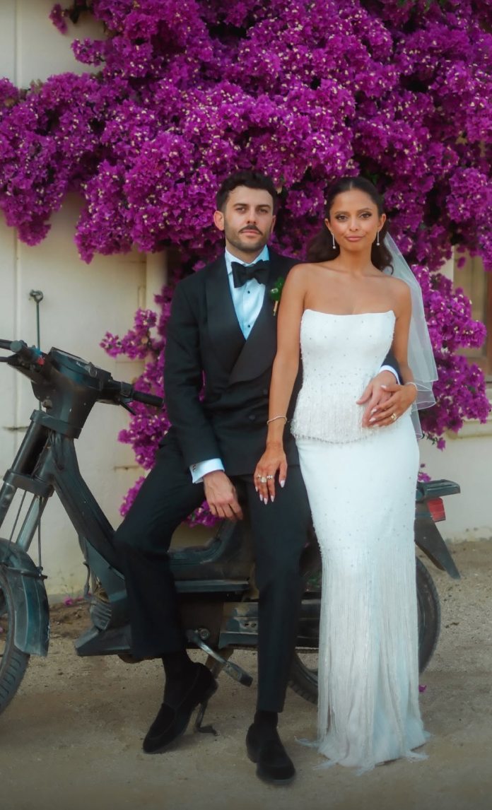 A bride and groom in wedding attire standing in front of a large purple flowering bush with a scooter nearby.