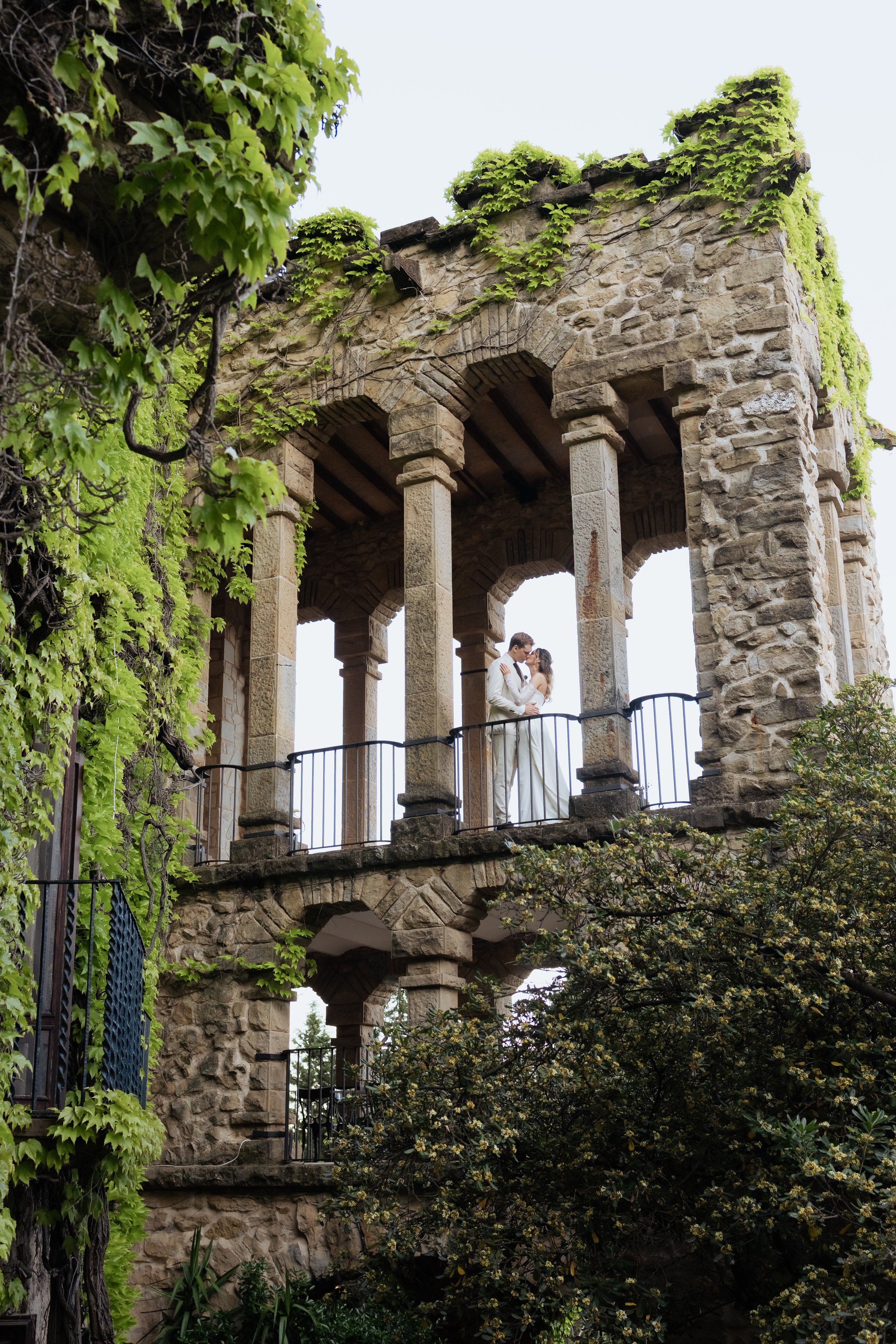 A couple dressed in wedding attire standing on a stone balcony of an old stone tower with ivy and greenery growing around it.