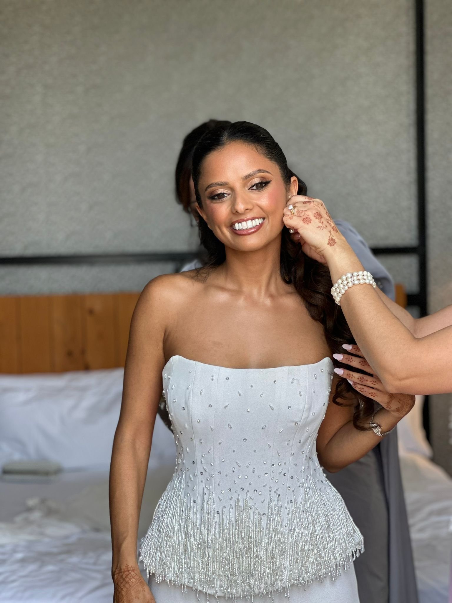 A smiling woman in a strapless white dress decorated with small beads, with henna on her hand and pearl bracelets, standing indoors with a gray wall and bed in the background.