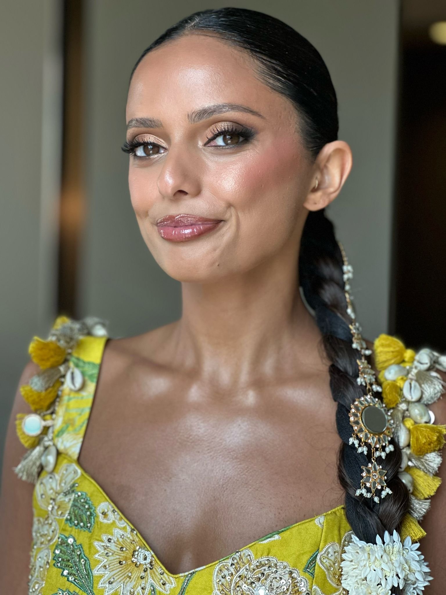 Close-up of a woman with dark braided hair, wearing a yellow dress with white embroidery and floral accents, smiling softly, with decorative jewelry.