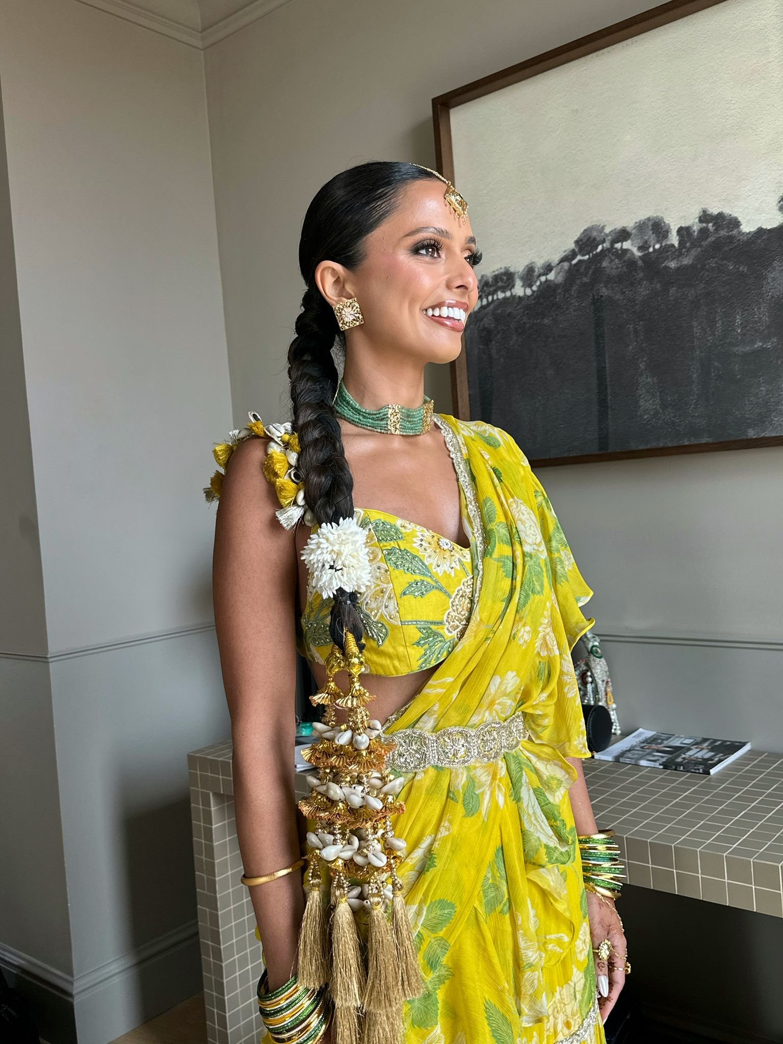A woman in a yellow saree with green leaf patterns, accessorized with traditional jewelry including earrings, headpiece, necklace, and bangles, standing indoors near a wall with artwork.