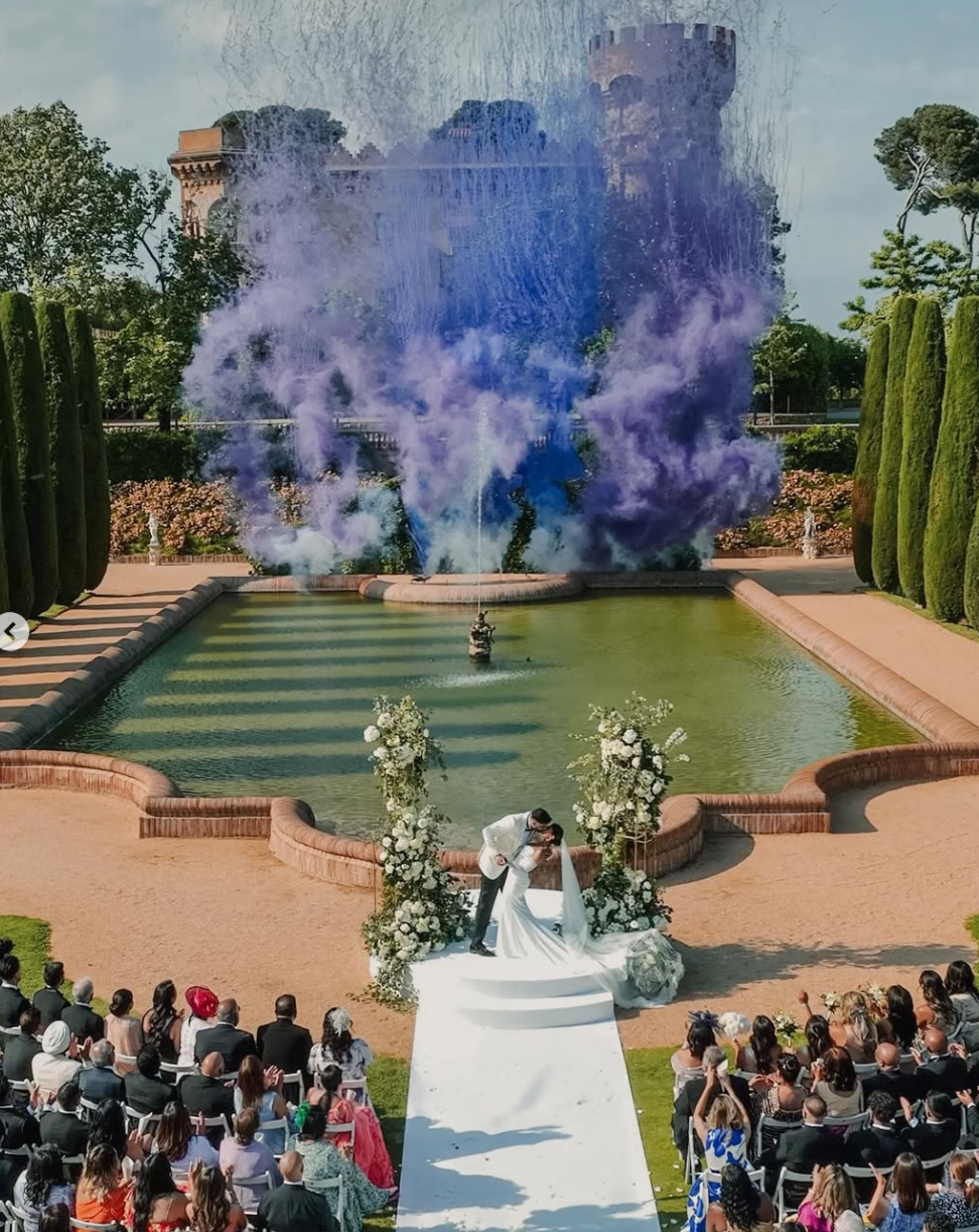 A wedding ceremony taking place outdoors with a bride and groom kissing in front of a fountain. The fountain is spraying purple powder in the air, creating a colorful explosion. Guests are seated around the ceremony, and the backdrop includes a large