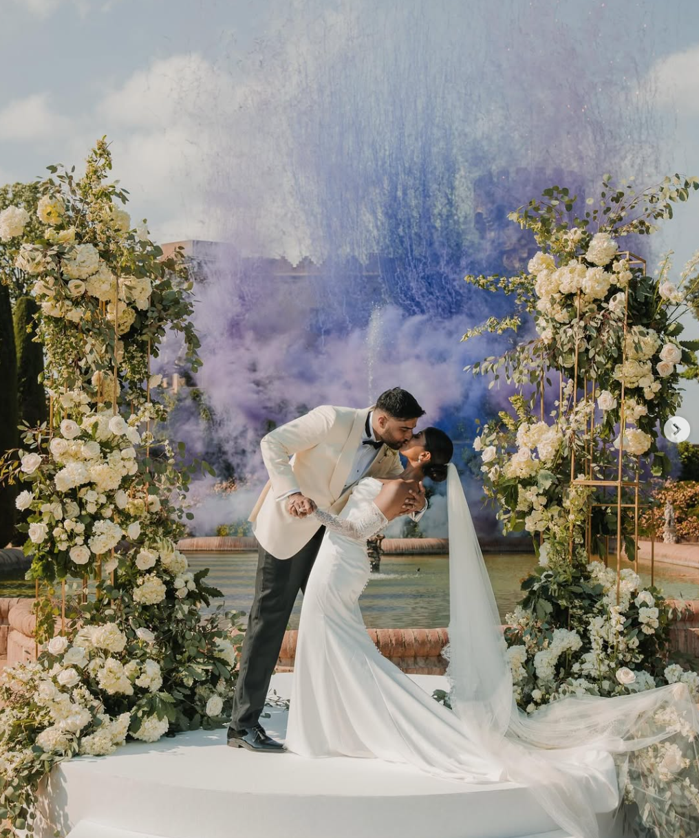Wedding couple kissing on a decorated platform outdoors, surrounded by white flowers and greenery, with a fountain in the background.