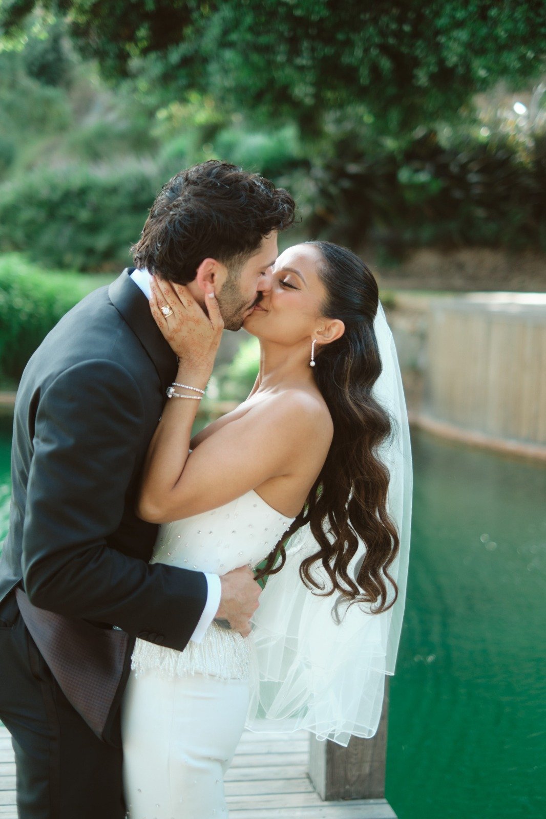 A bride and groom kissing outdoors near a pond, with trees and greenery in the background.