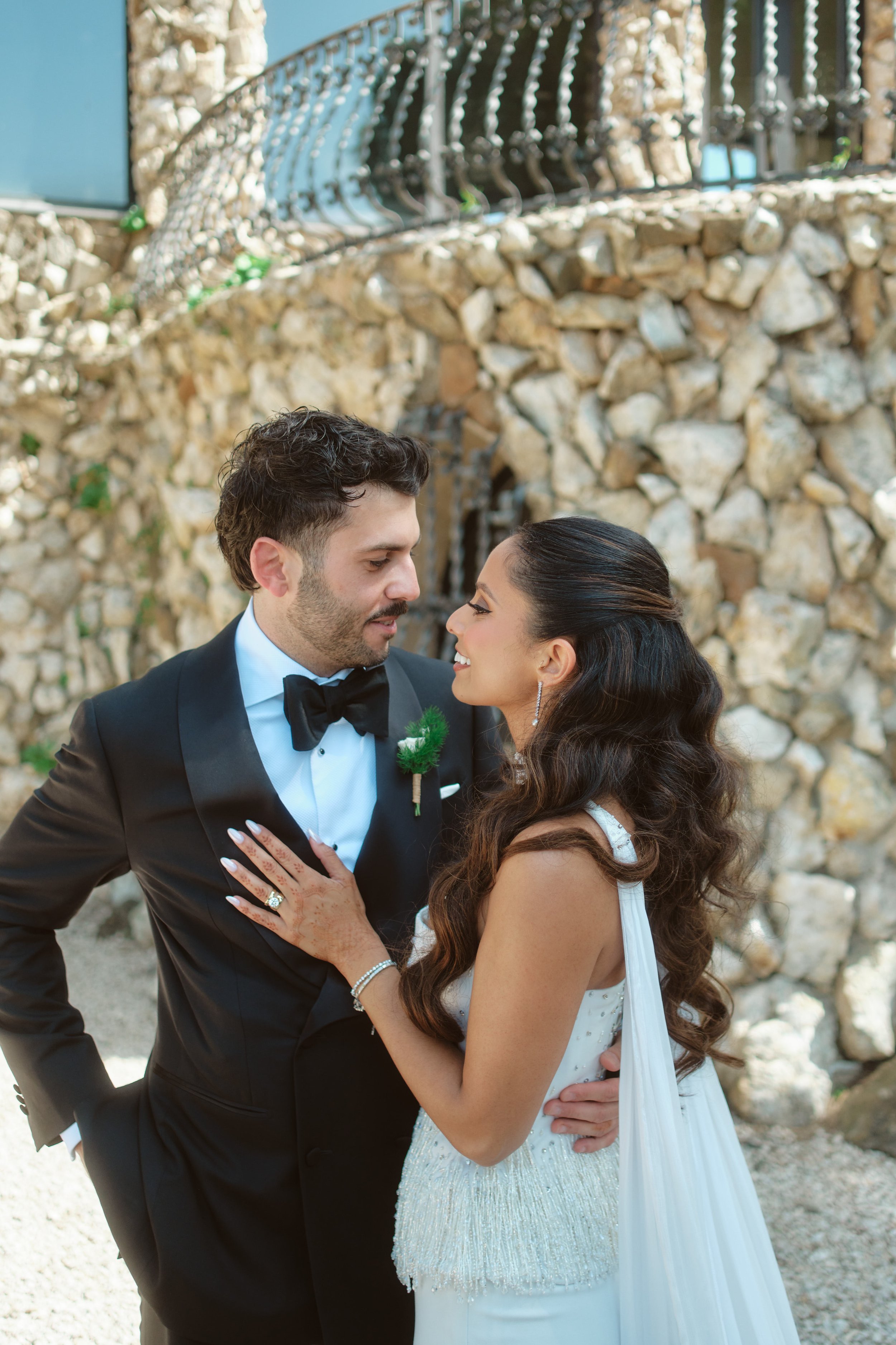 A bride and groom are close together outdoors, smiling at each other. The groom wears a black tuxedo and bow tie, while the bride wears a white sleeveless wedding gown with beading and jewelry. The background features a stone wall and a metal railing