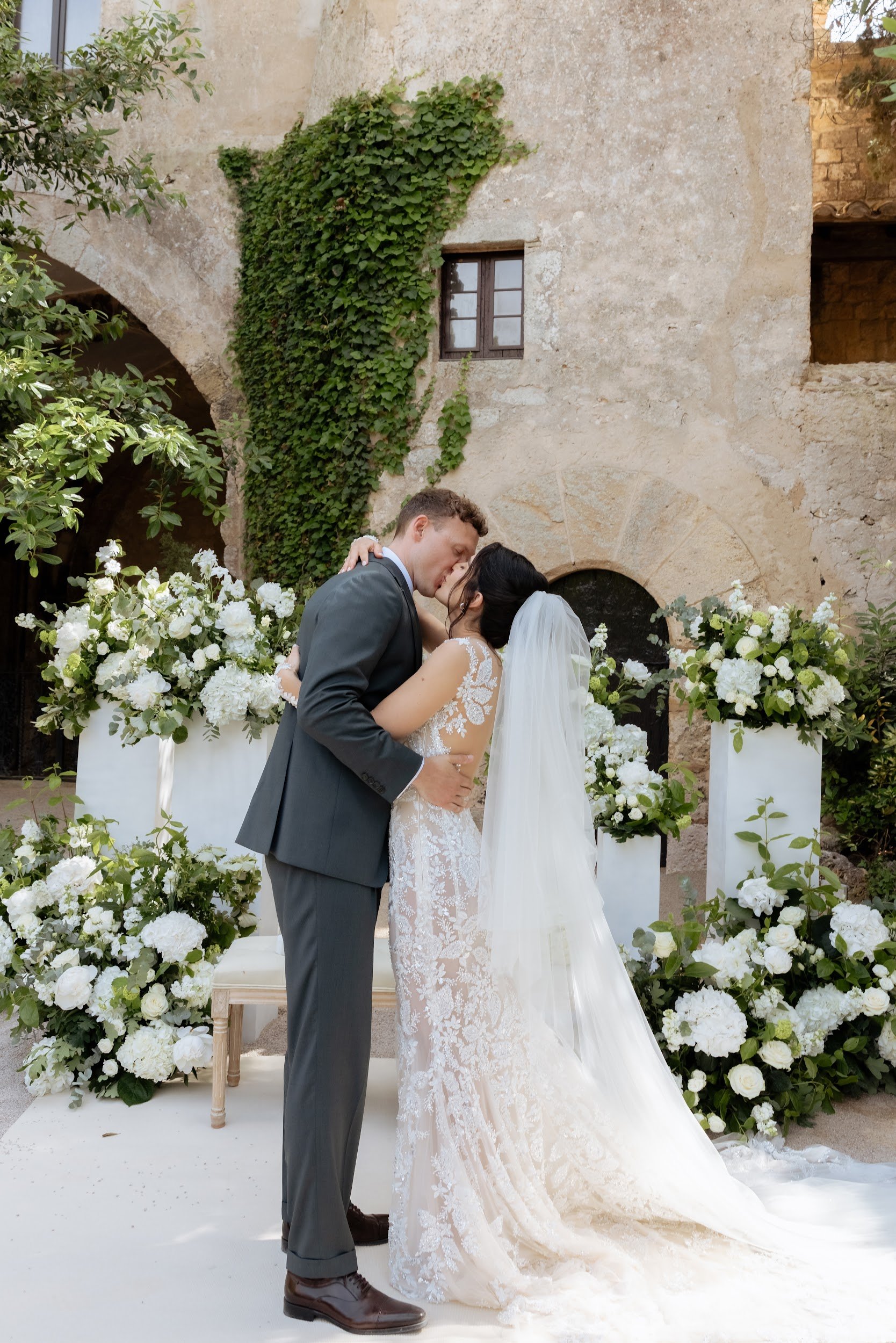 A bride and groom share a kiss during their wedding ceremony, surrounded by white floral arrangements and greenery, with a rustic stone building in the background.